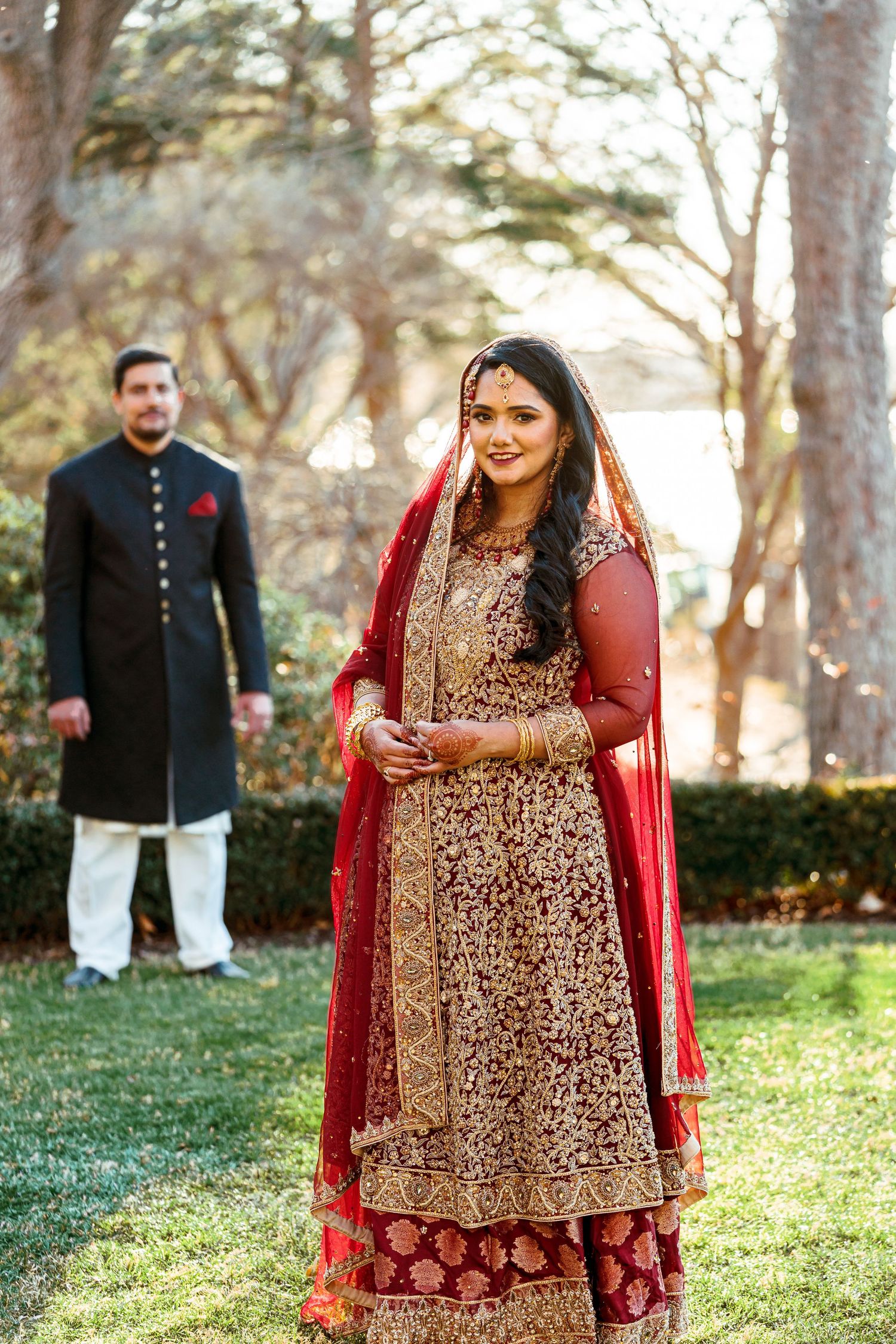 A couple in traditional South Asian wedding attire poses in a garden setting with intricate embroidered outfits.