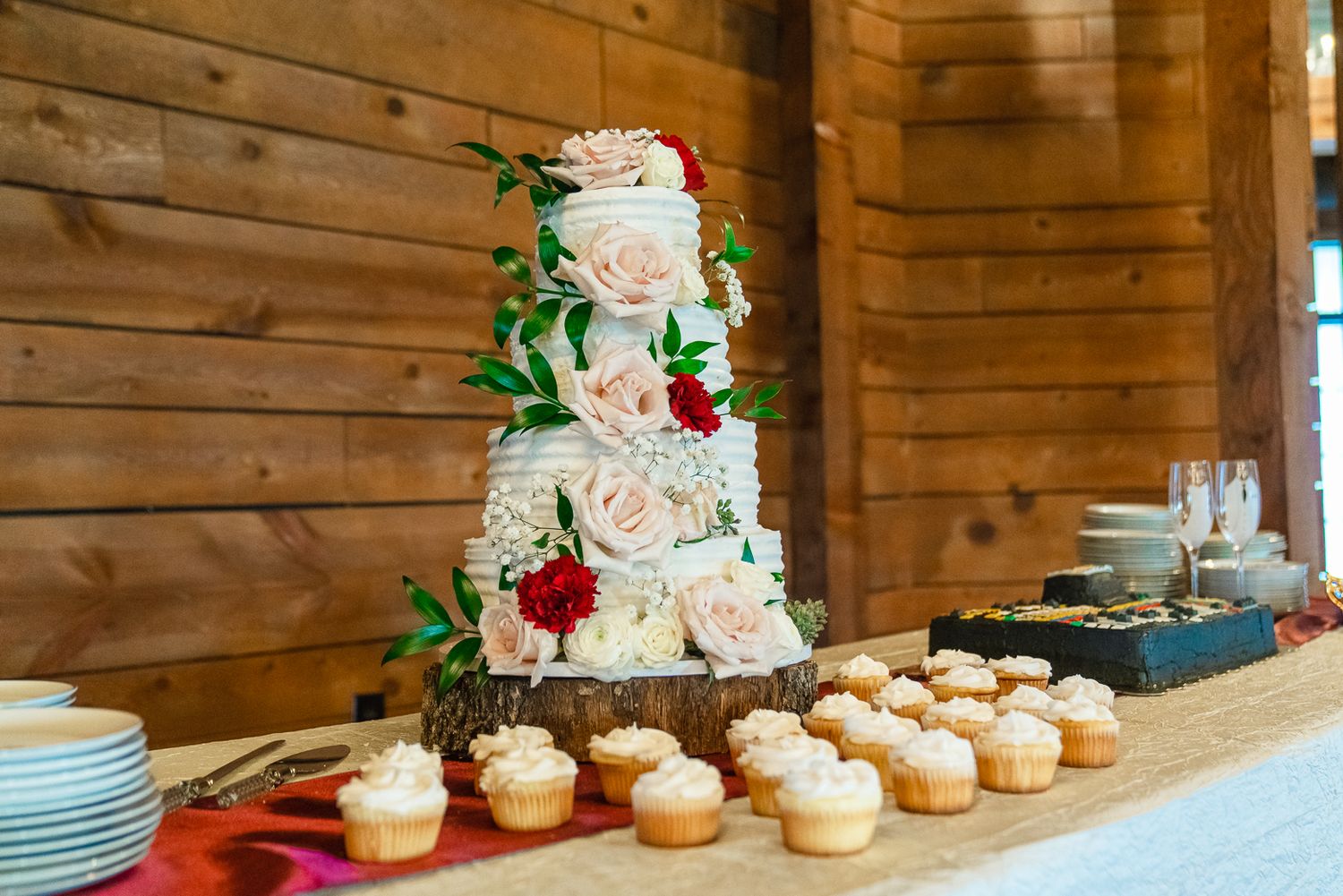 Rustic wooden dessert display with elegant white wedding cake and cupcakes decorated with pink and red roses.