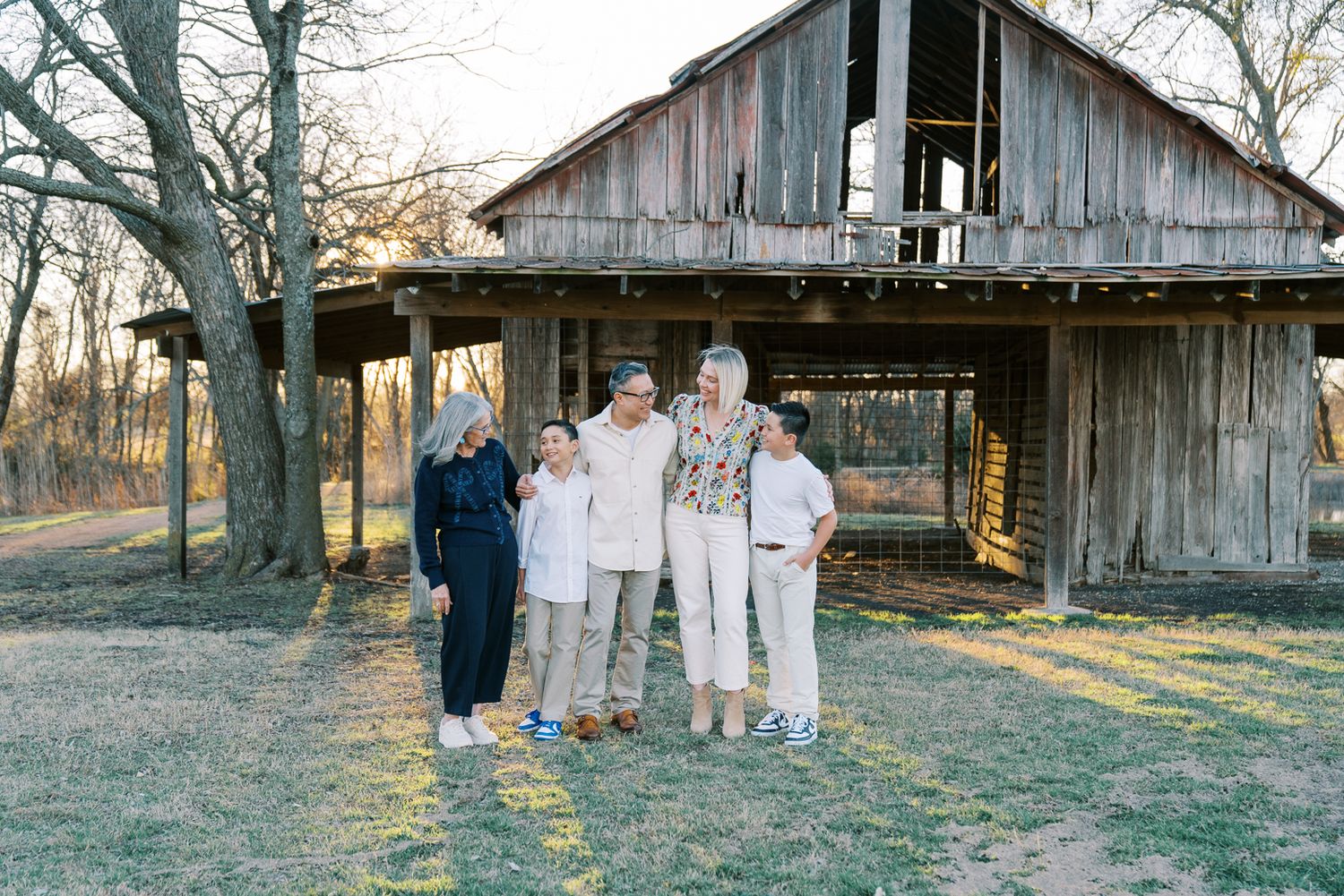 A rustic wooden barn with peeling paint stands in a field during golden hour as people gather nearby.