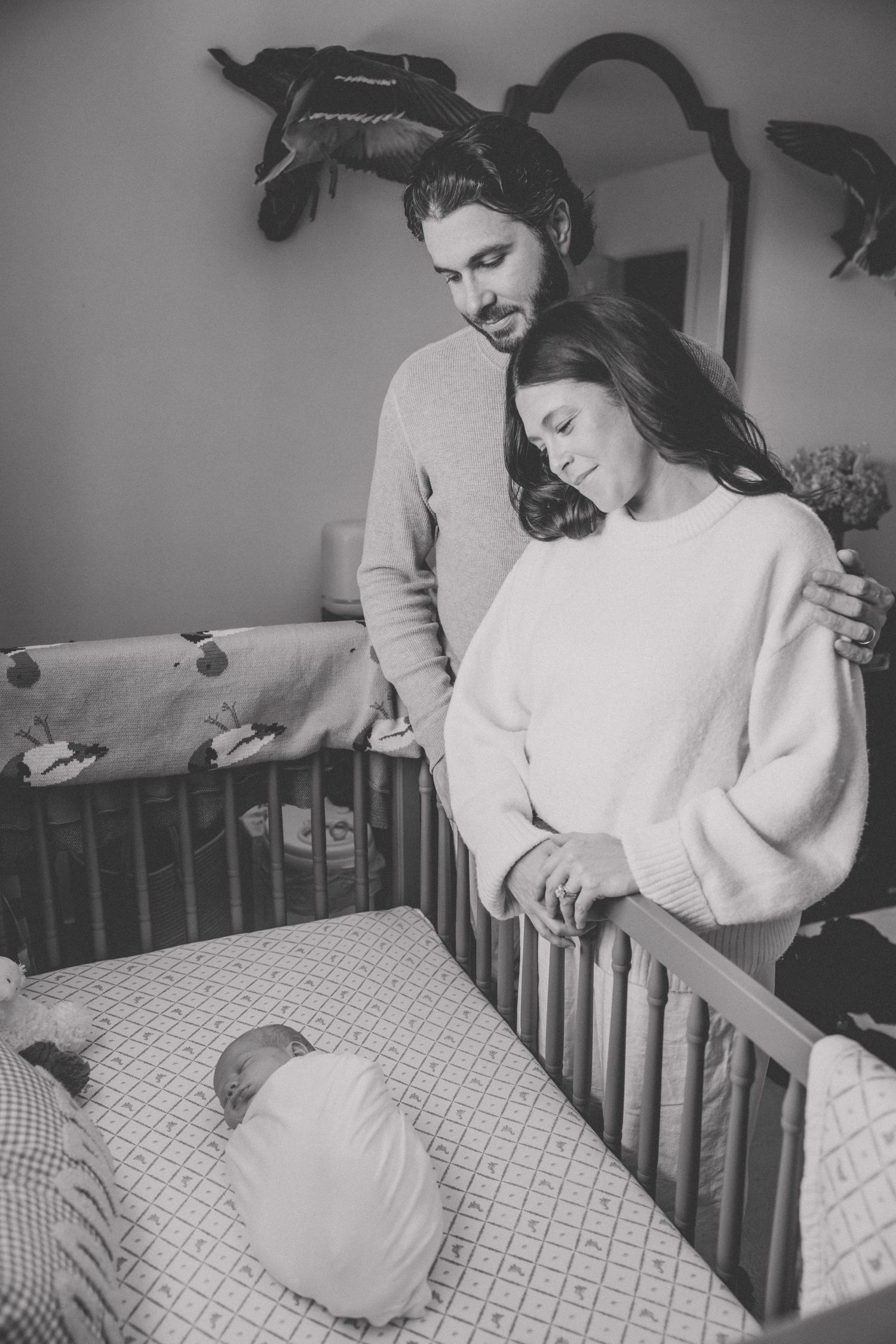 Black and white photo of couple lovingly gazing at newborn baby lying in wooden crib in nursery room.