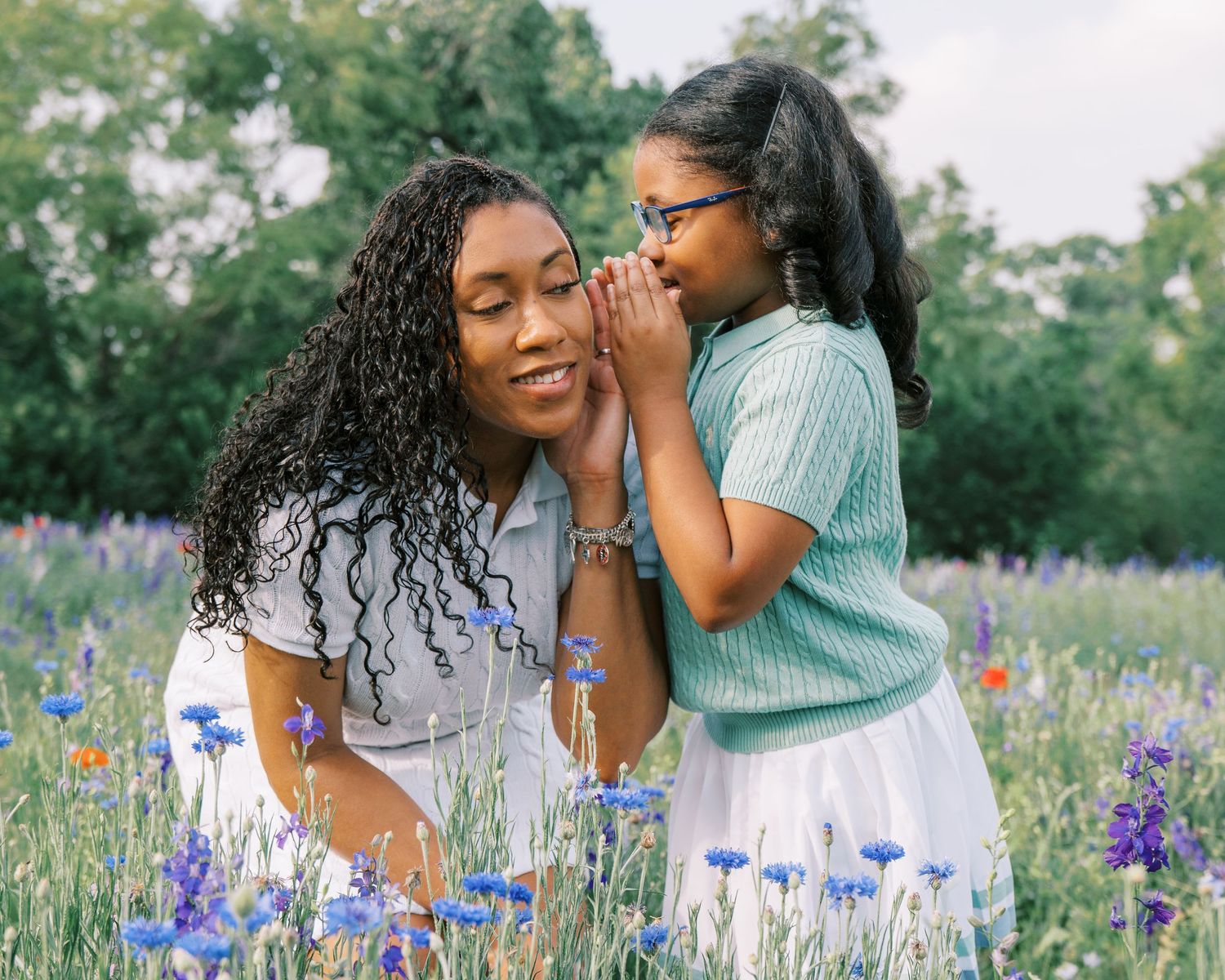A mother and daughter share a sweet whisper moment in a field of blue wildflowers on a sunny day.