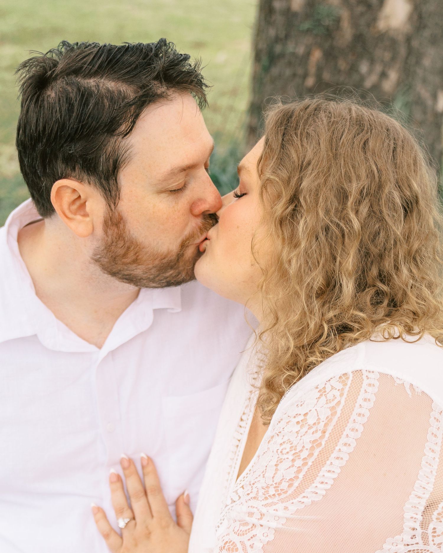 Couple sharing a romantic kiss outdoors, woman wearing white lace dress with engagement ring visible.