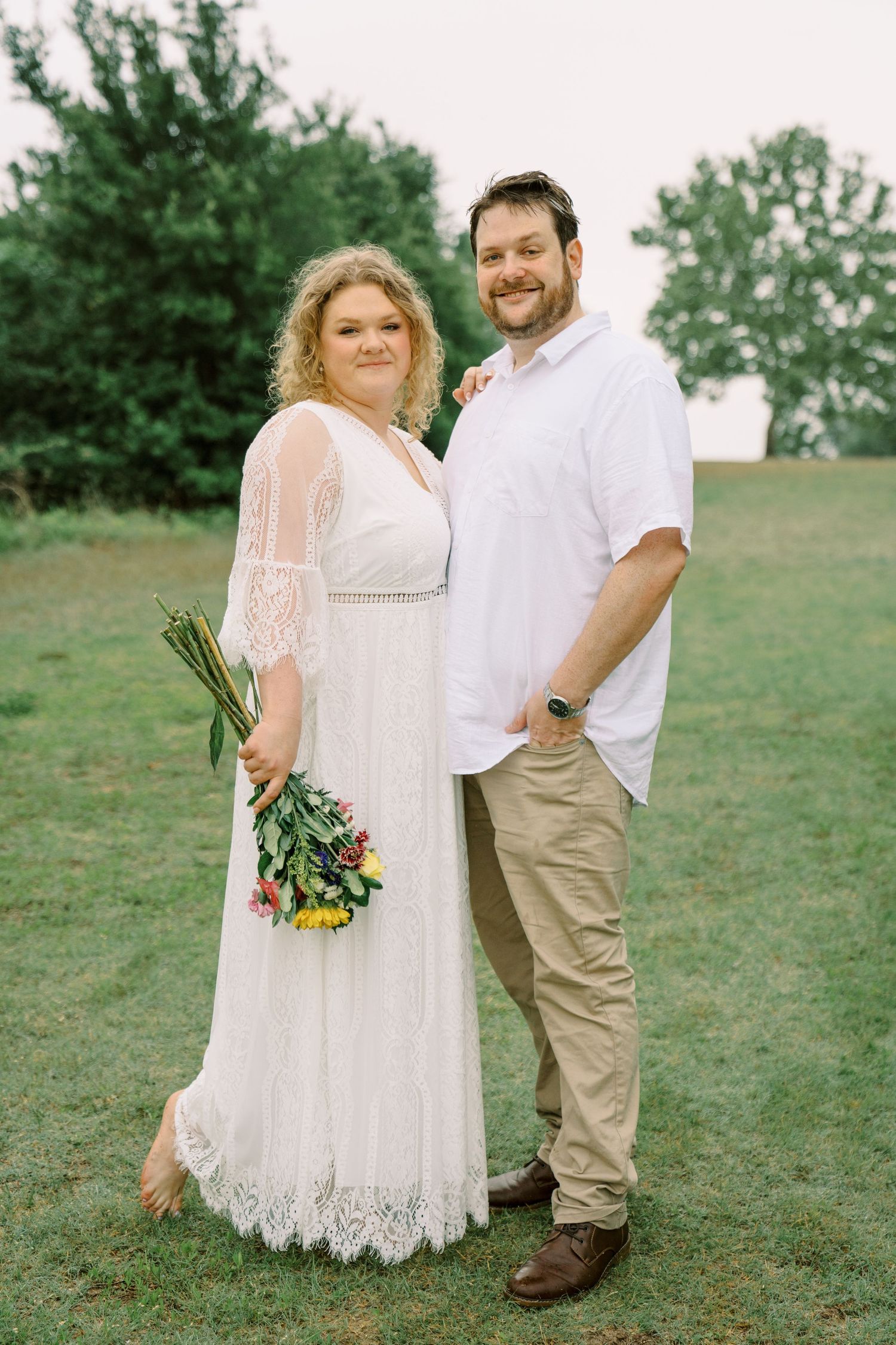 Bride in white lace dress holding colorful bouquet poses with groom in white shirt on green lawn.