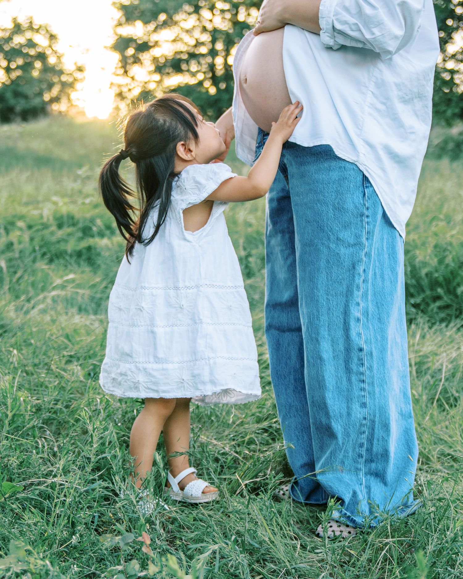 Young girl in white dress kissing pregnant mother's belly in a sunny green field at golden hour.