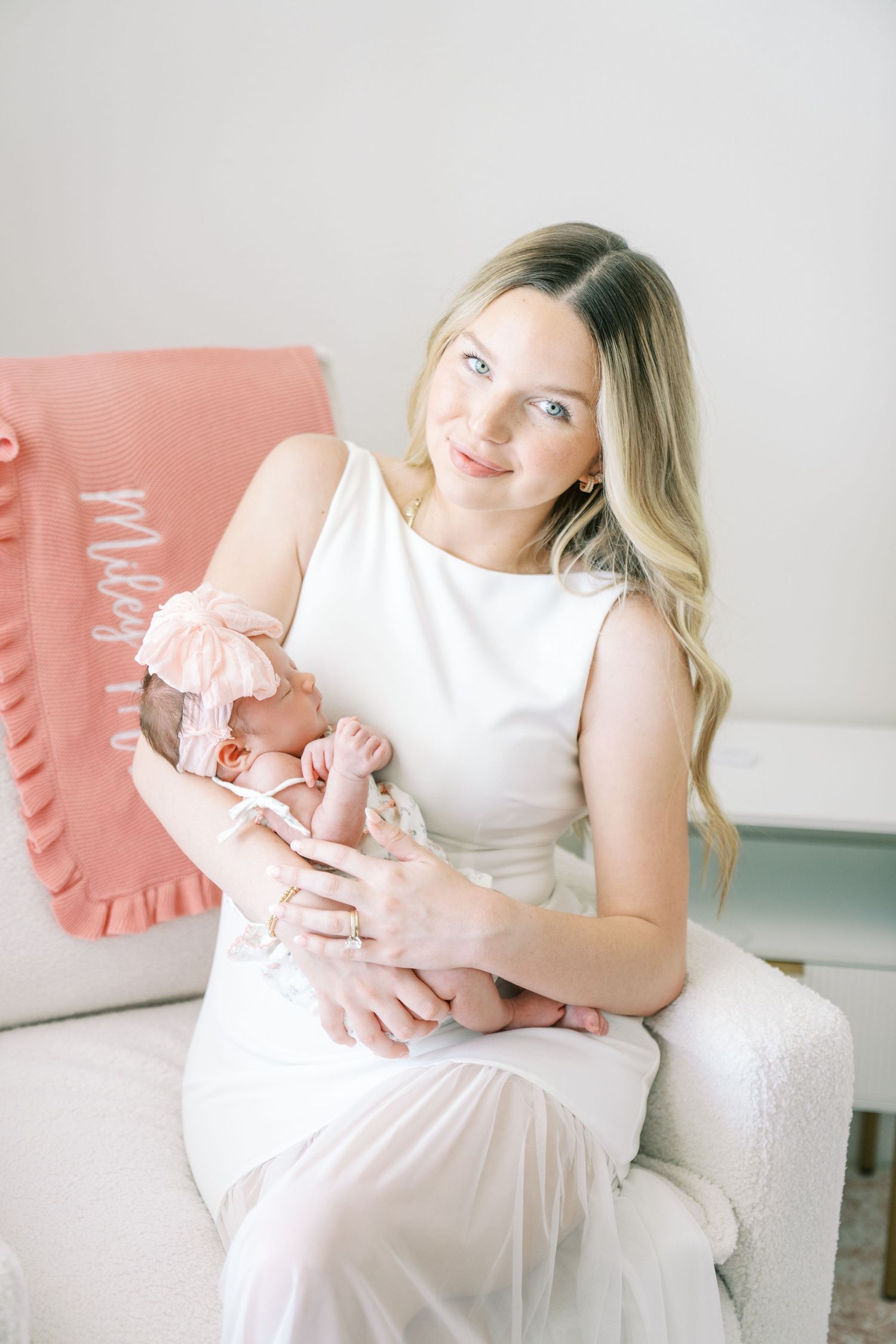 A smiling young mother in a white dress holds her sleeping newborn baby dressed in pink in a bright, airy nursery.