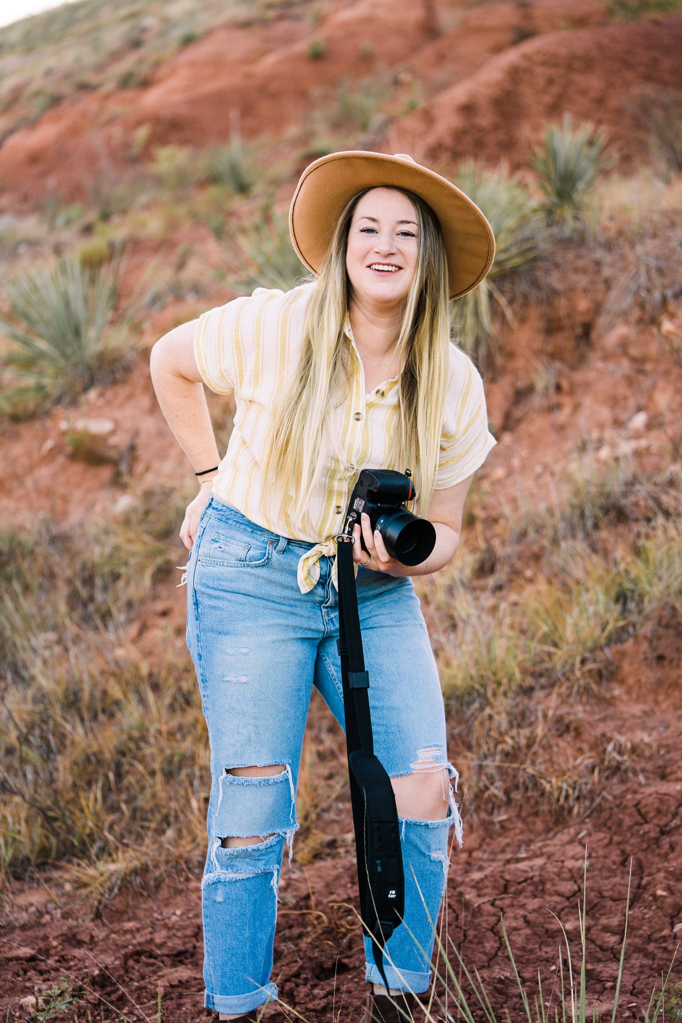 A photographer in a wide brim hat and ripped jeans poses with a camera against red rock formations.