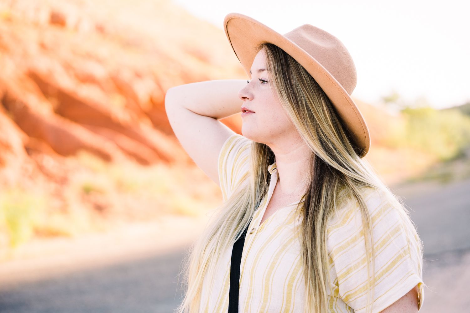 Person in a wide brim hat and casual attire poses against scenic red rock formations at sunset.