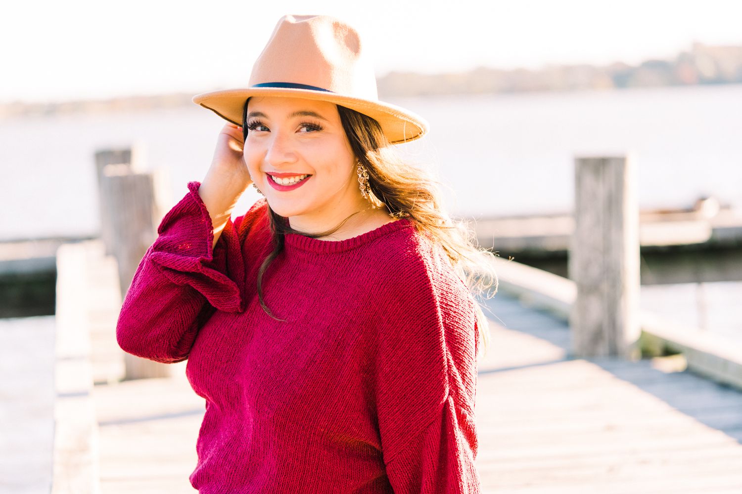 A person in a burgundy sweater and beige hat stands smiling on a dock by the water on a sunny day.