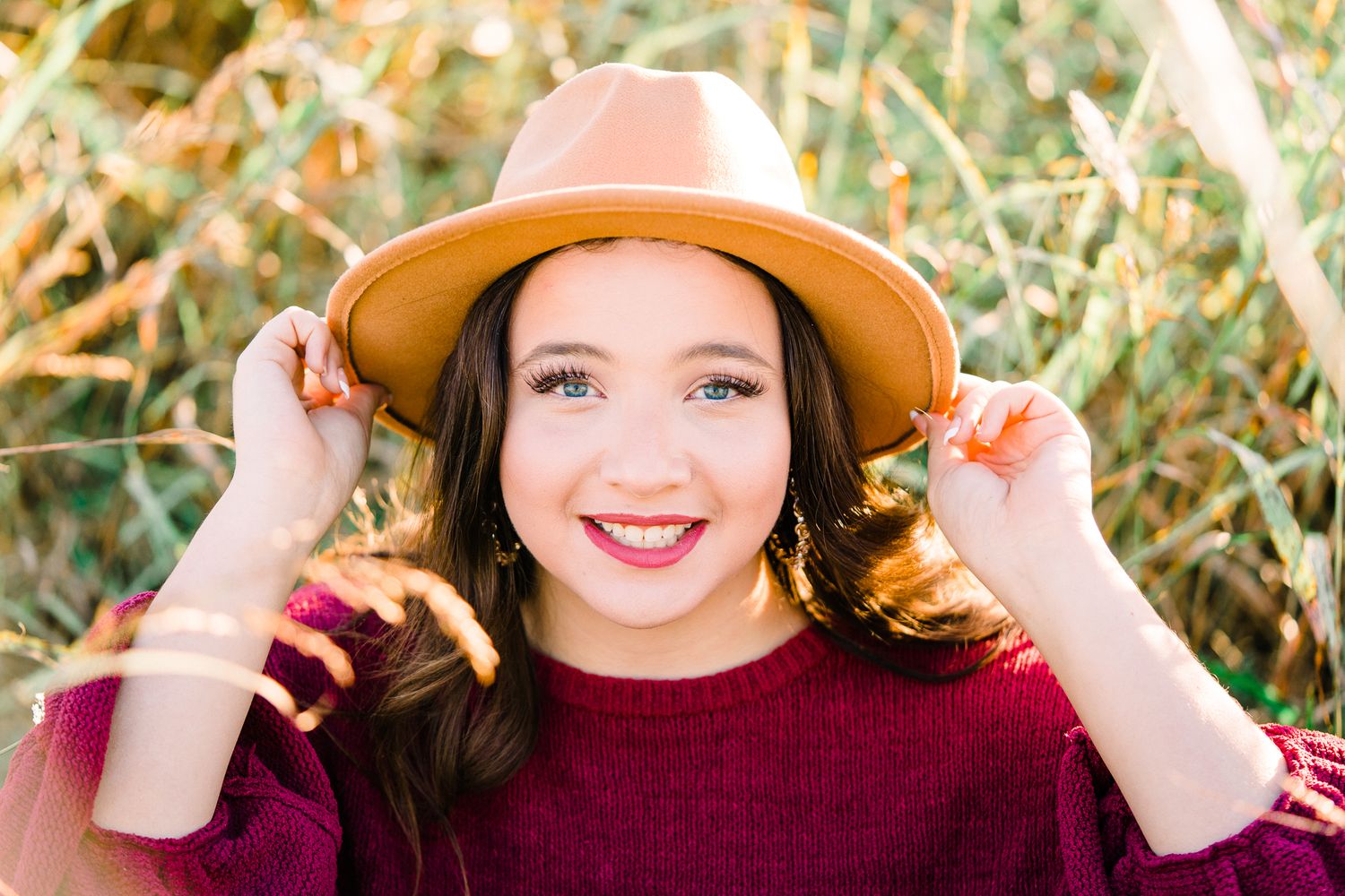 A person wearing a burgundy sweater and brown hat poses outdoors in autumn sunlight.