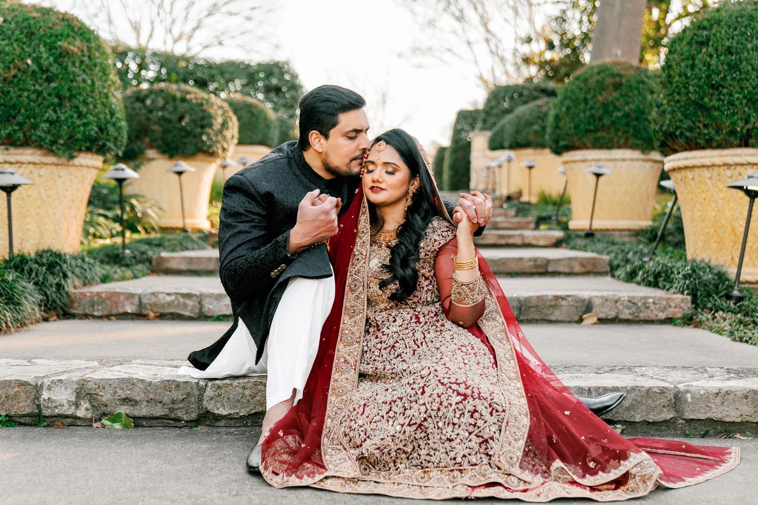 A couple in ornate Indian wedding attire shares playful moments on stone steps outdoors.
