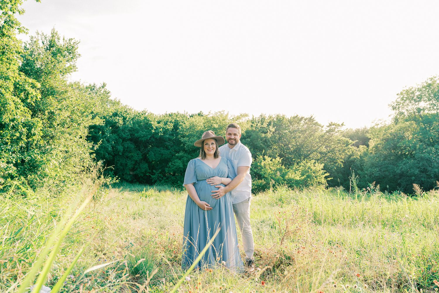 A couple embraces in a sunlit meadow with tall grass and wildflowers during a summer photo session.