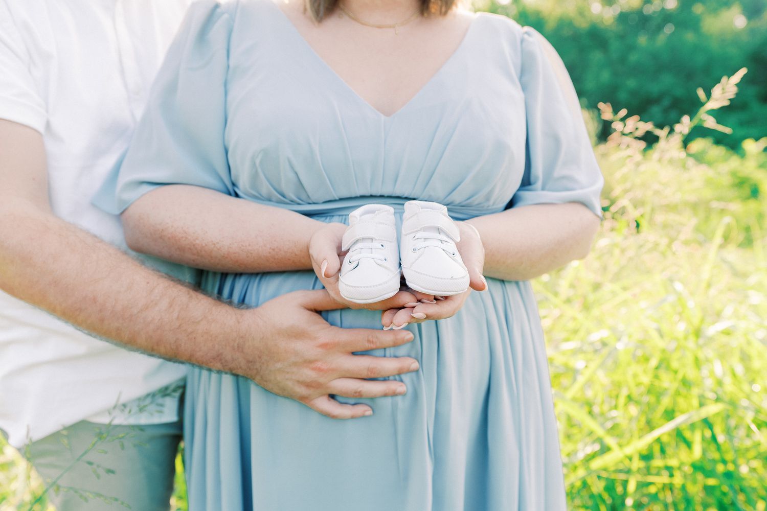 A person in a light blue maternity dress stands in a sunny wildflower meadow holding their baby bump.