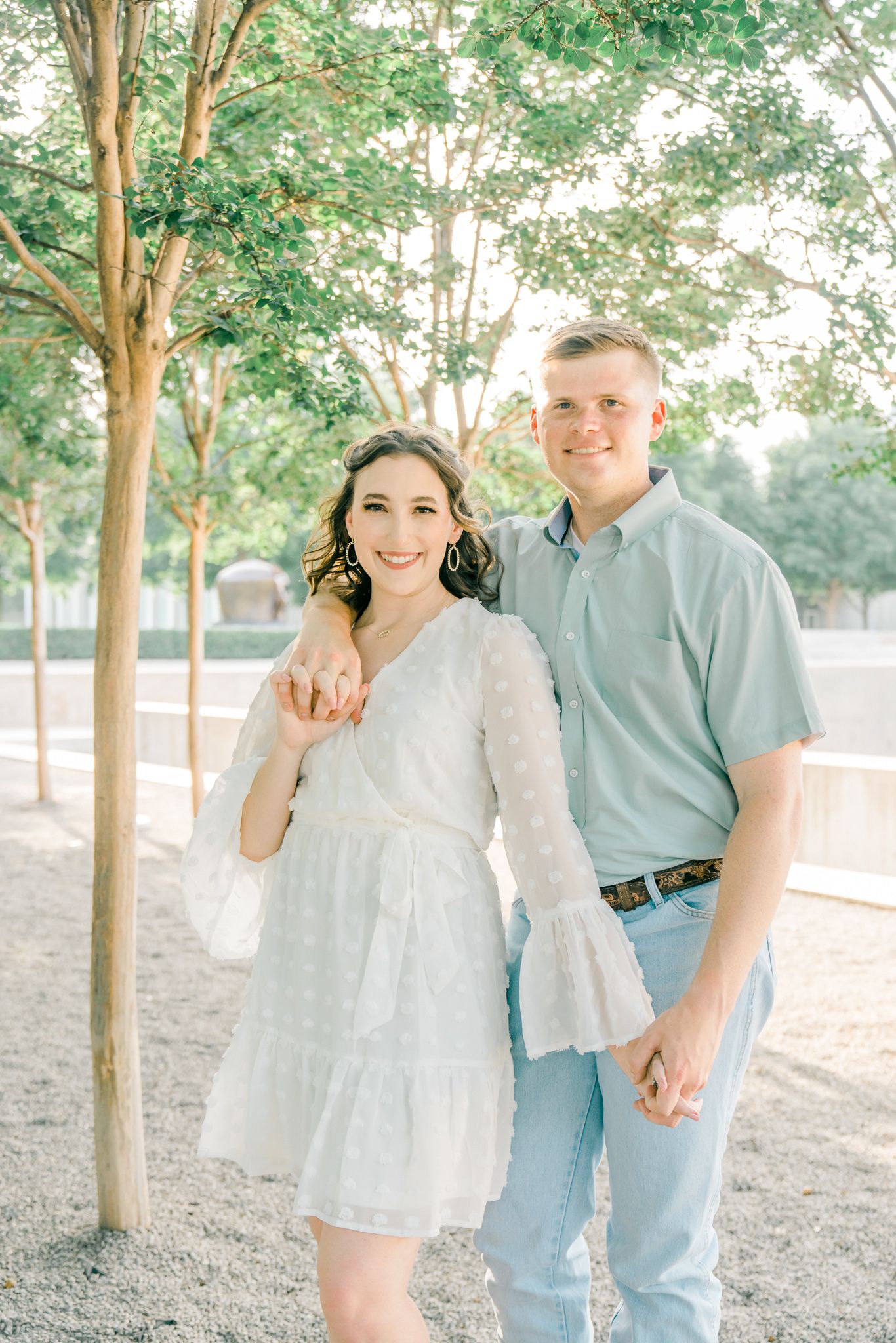 A couple in pastel outfits poses playfully among trees during a sunny outdoor photoshoot.