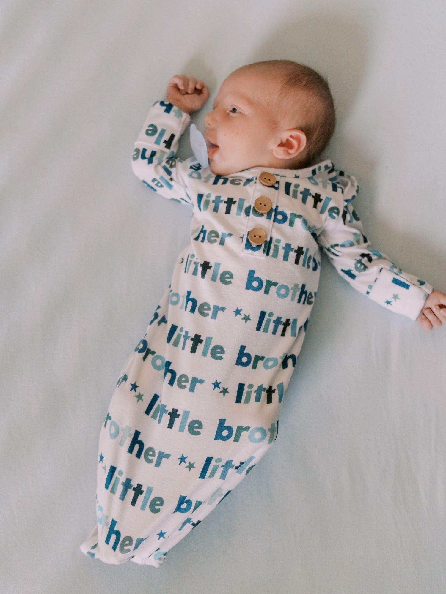 A sleeping baby wearing a white onesie with repeating 'little brother' text pattern lies on a gray background.