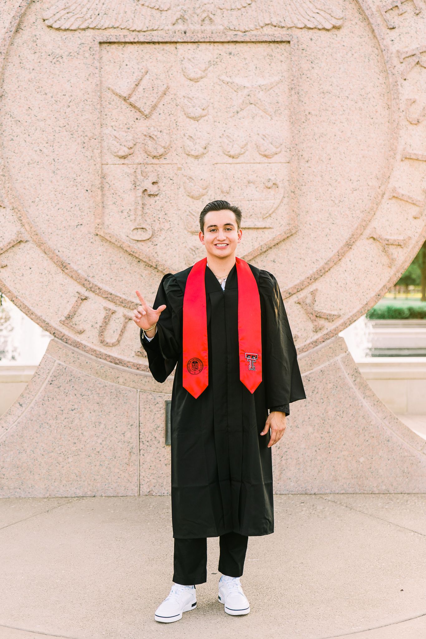 Graduate in black cap and gown with red stole poses in front of stone wall during graduation photo session.