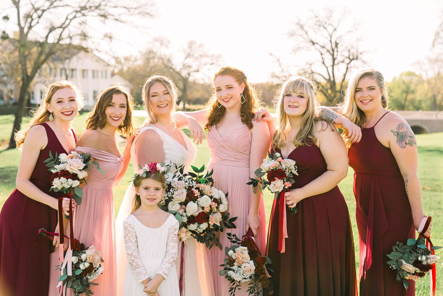 Wedding party in pink and burgundy dresses poses together outdoors holding floral bouquets during sunset.