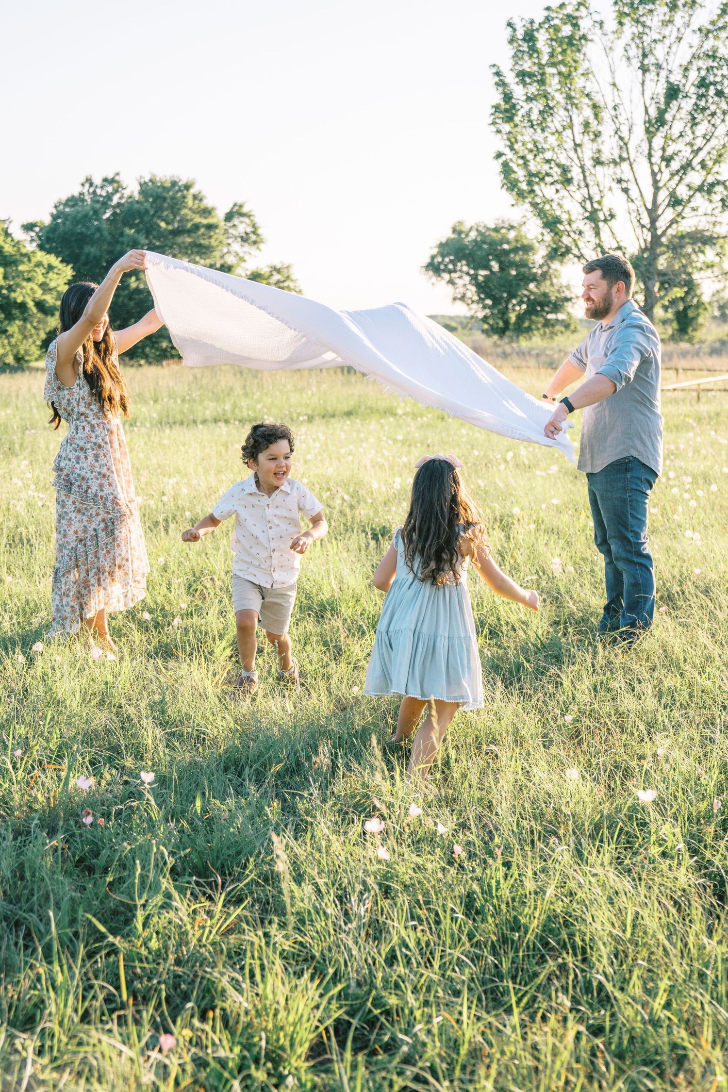 Family of four playing with a white blanket in a sunny green meadow, parents lifting it over two young children.