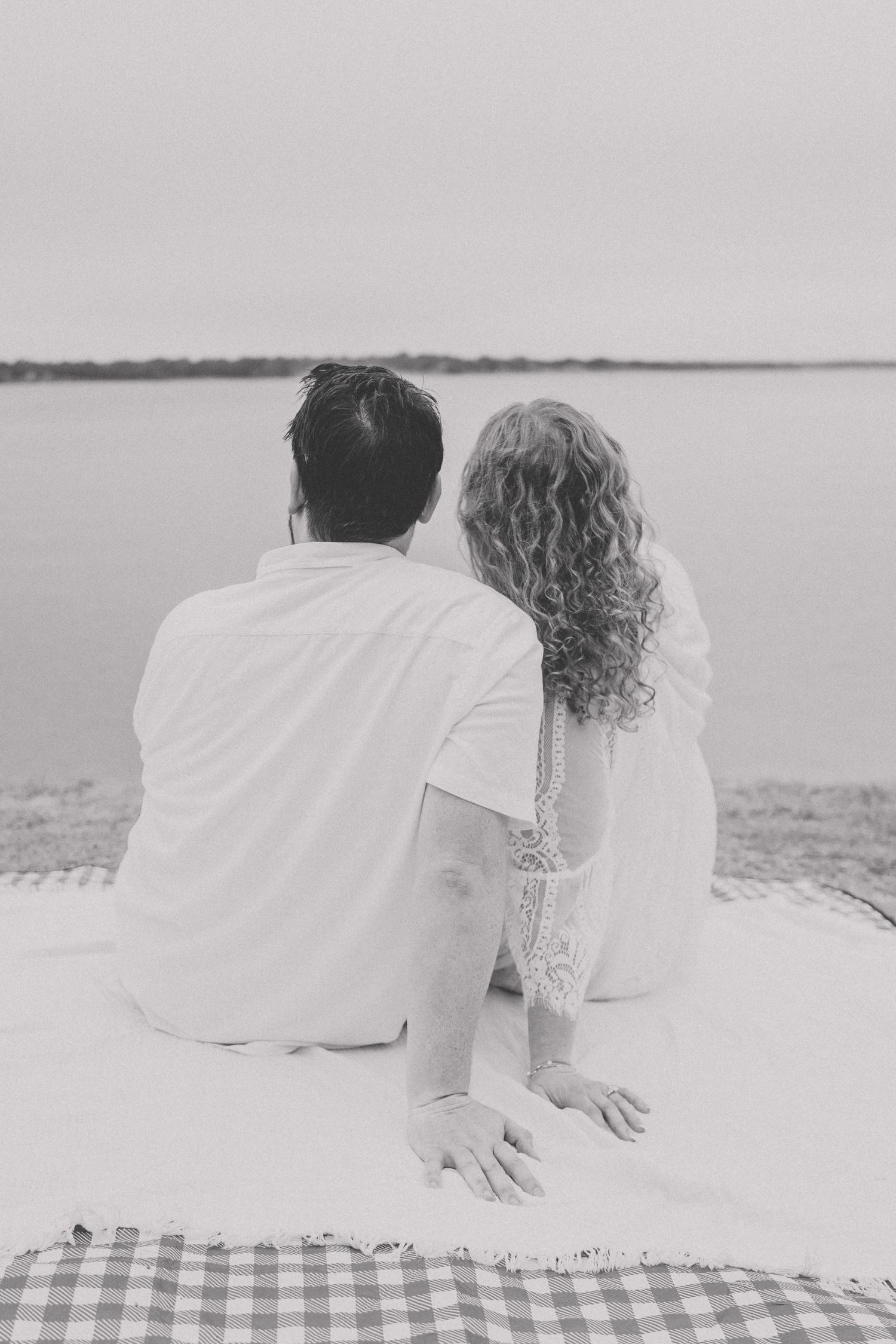 Black and white photo of couple sitting together on blanket gazing at calm lake water from behind.