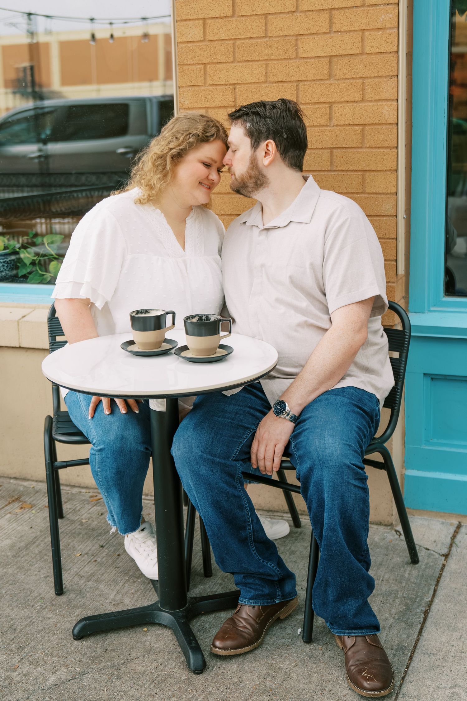 An engaged couple leans in close at an outdoor cafe table with two coffee cups between them.