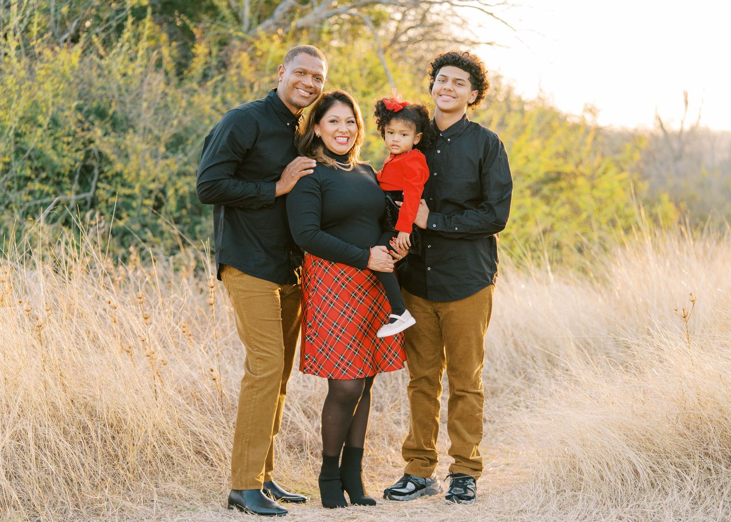 Family group poses together in casual outfits against backdrop of tall grass during golden hour.
