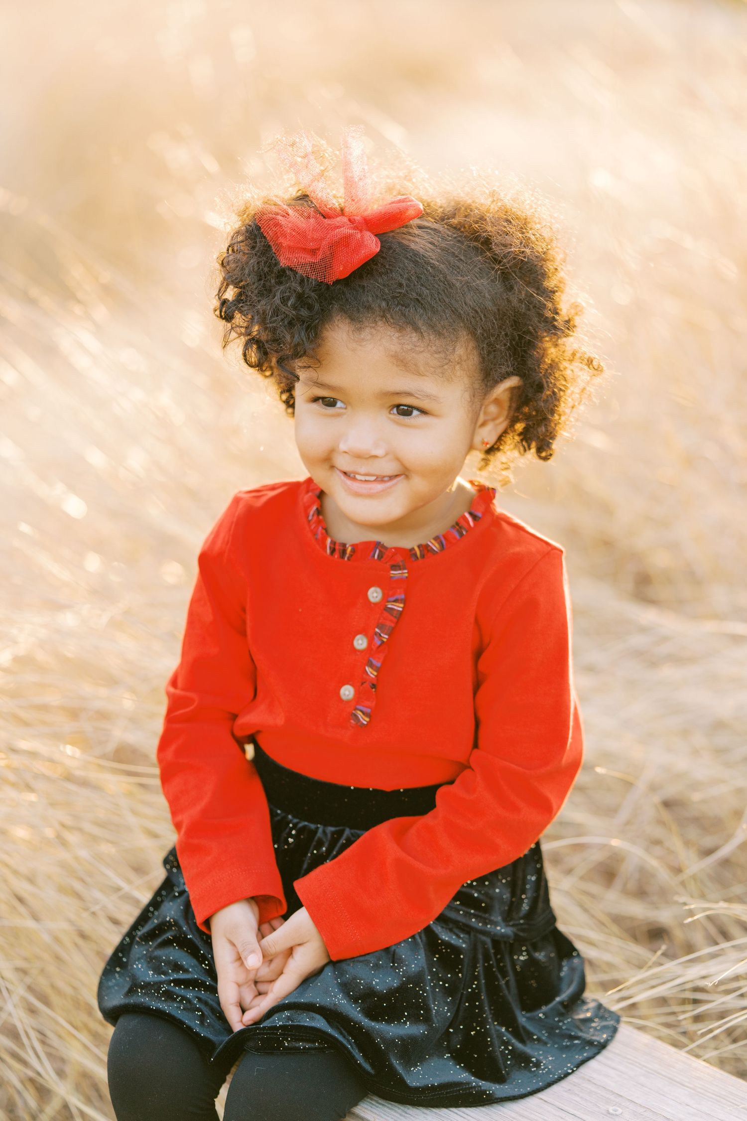 Young child wearing a bright red jacket and navy skirt sits in golden grass during autumn.