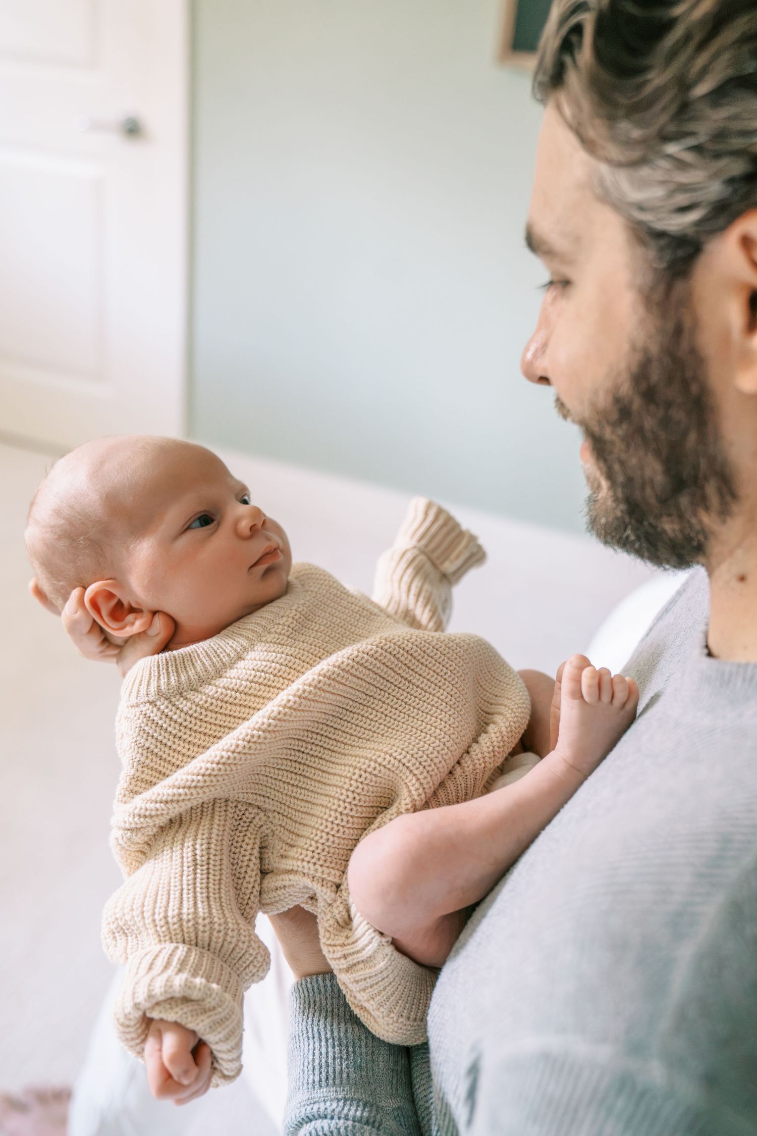 Bearded father tenderly holding his newborn baby dressed in a cream knit sweater indoors.