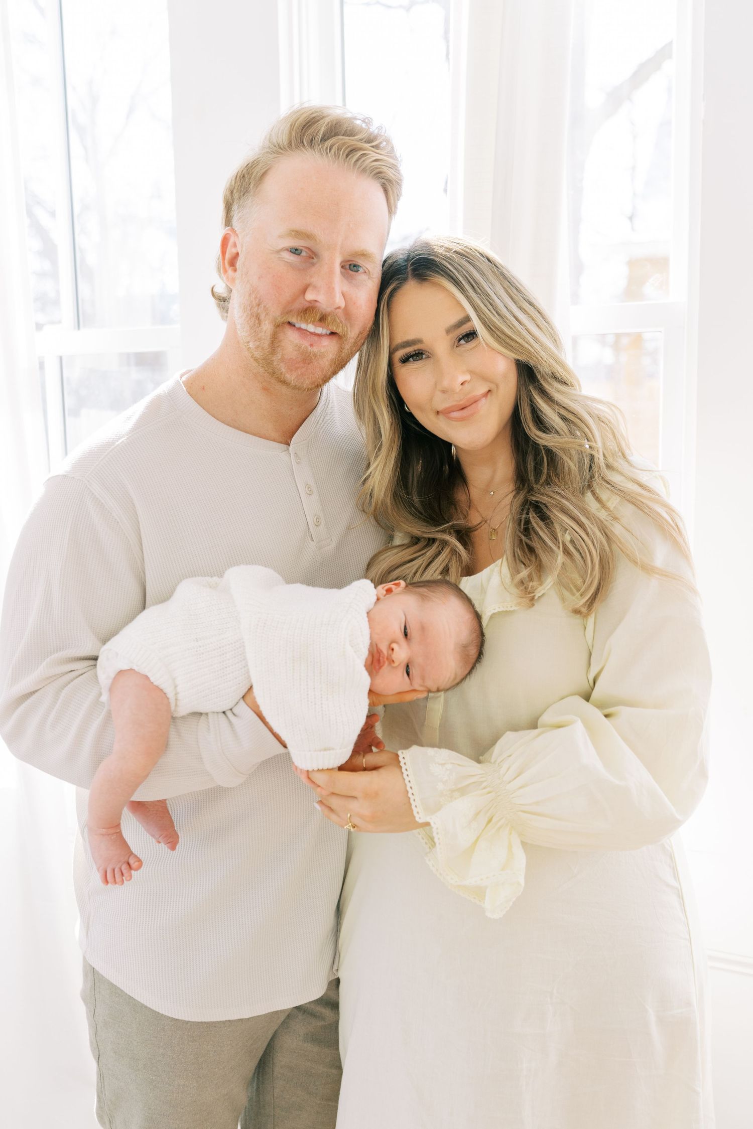 Family portrait in soft white clothing against sheer curtains and natural window light.