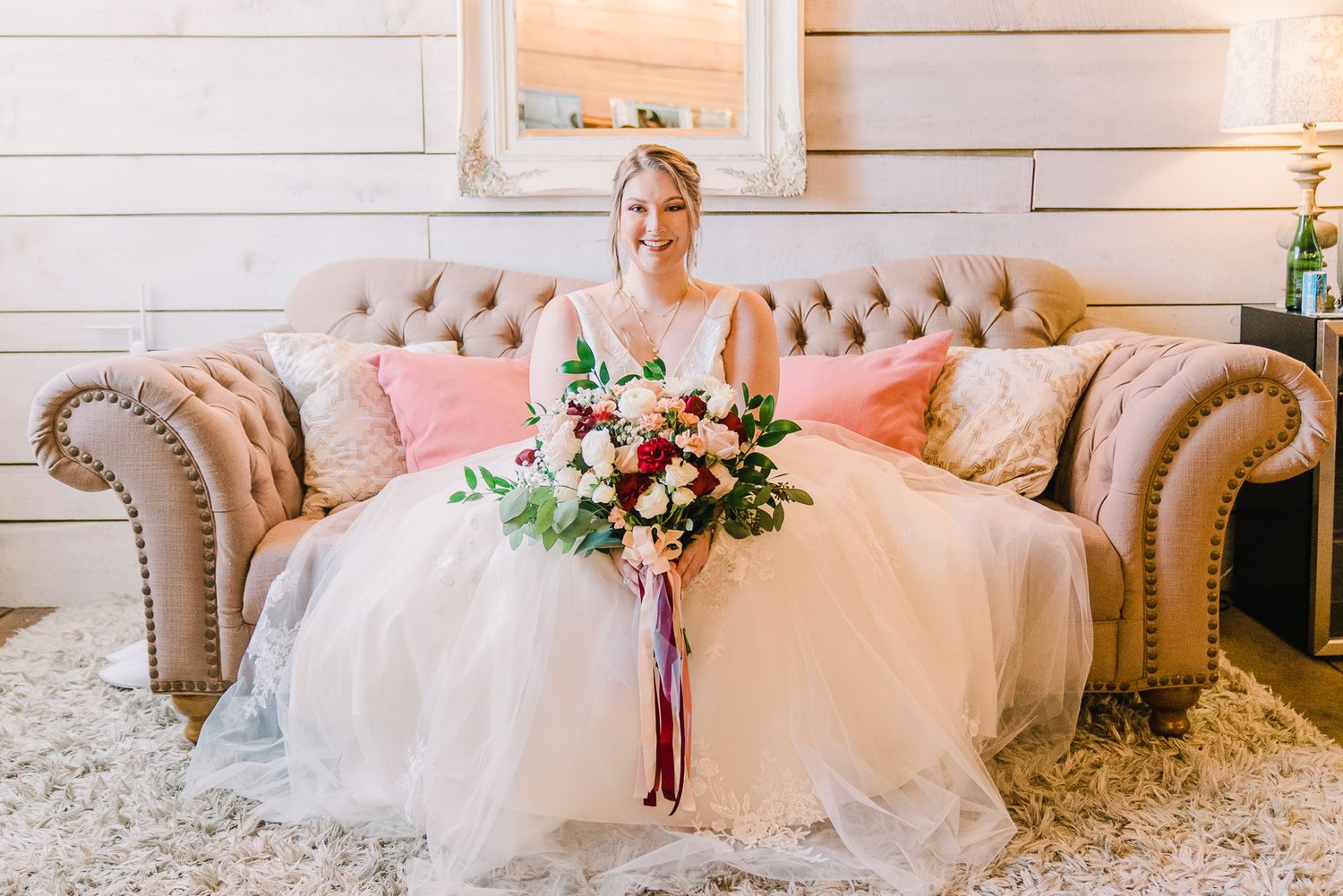 A bride in a white wedding gown sits on an elegant beige tufted sofa holding a pink and white floral bouquet.