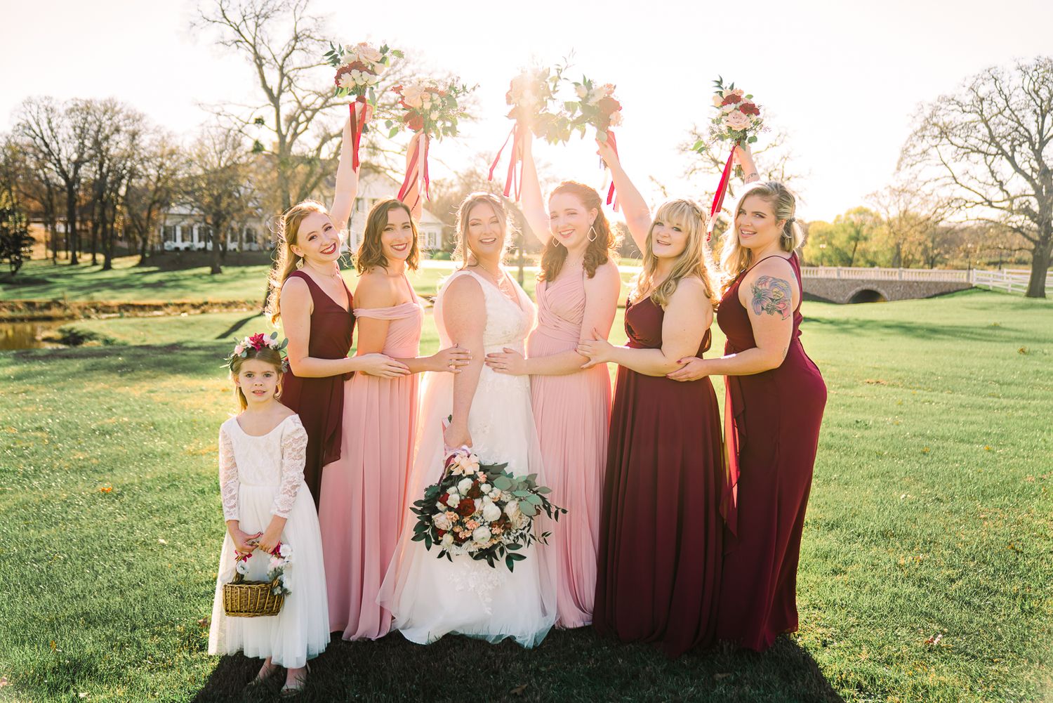 A group in formal attire stands together in a sunlit garden setting for a wedding photo session.