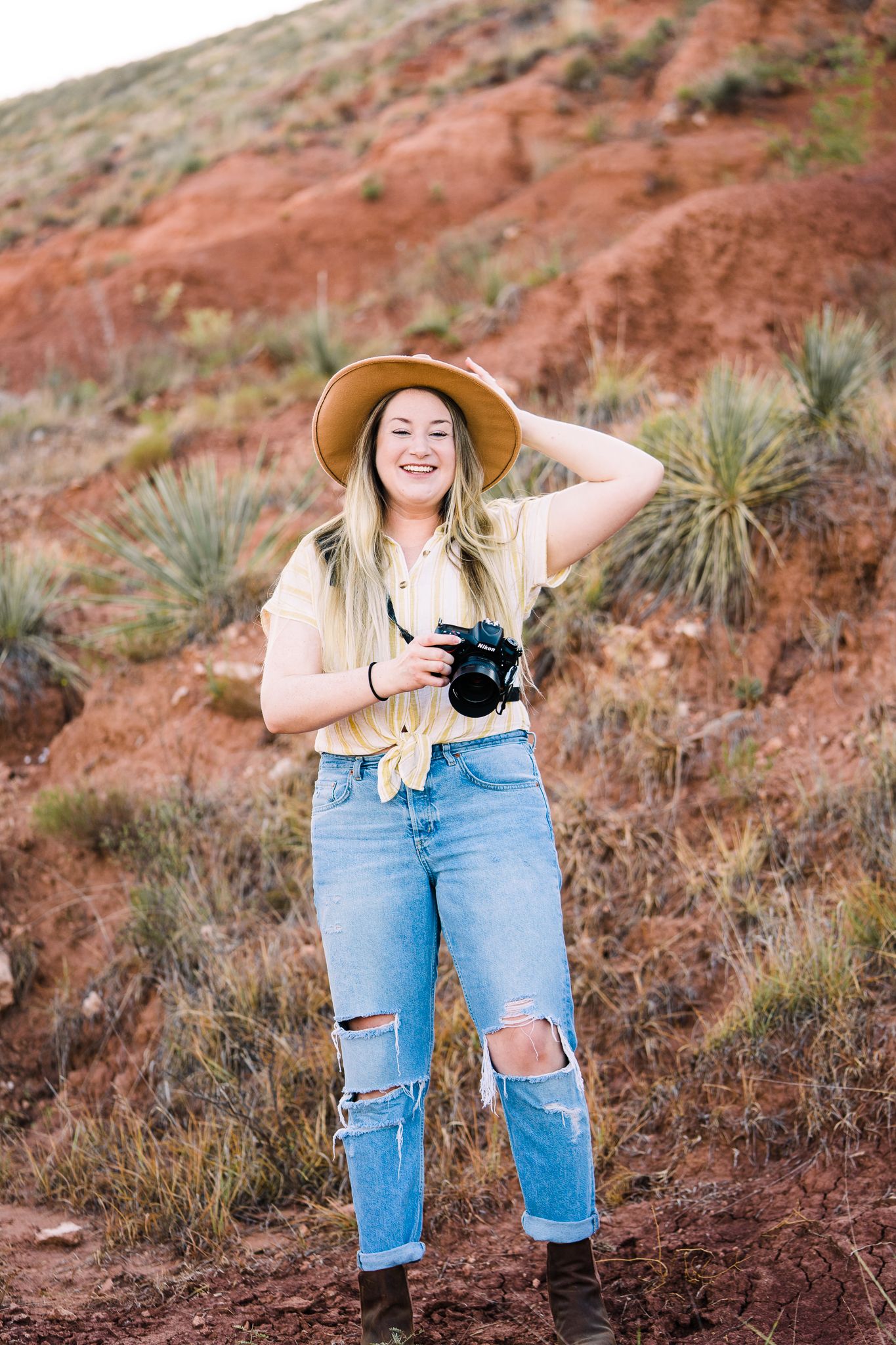 Photographer in ripped jeans and wide-brimmed hat explores red rock terrain with camera in hand.
