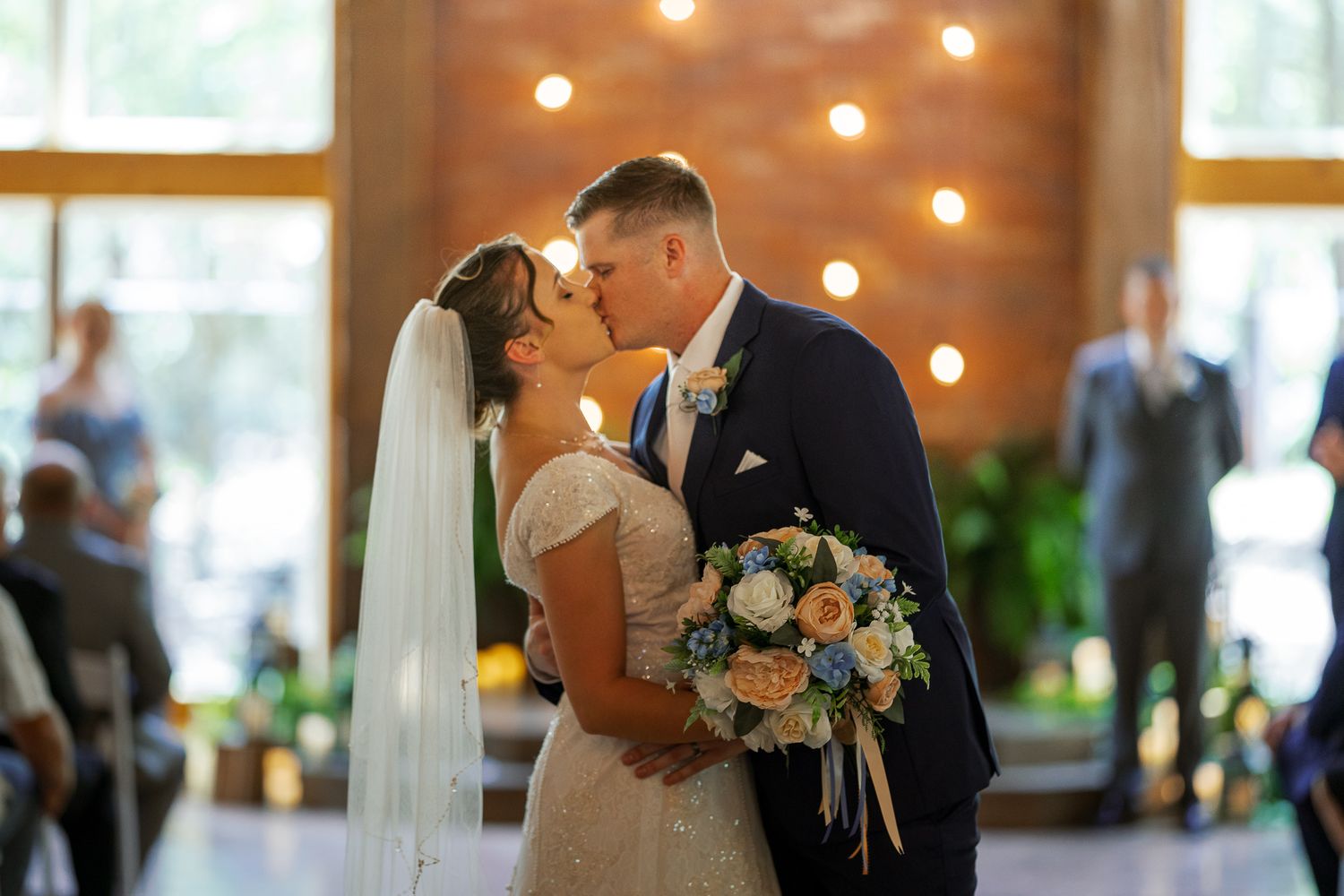 A couple shares a romantic kiss during their wedding ceremony in an elegant venue with twinkling lights and floral decor.