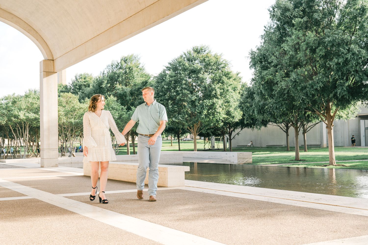 A couple walks together under an elegant white archway in a serene park setting during a summer day.