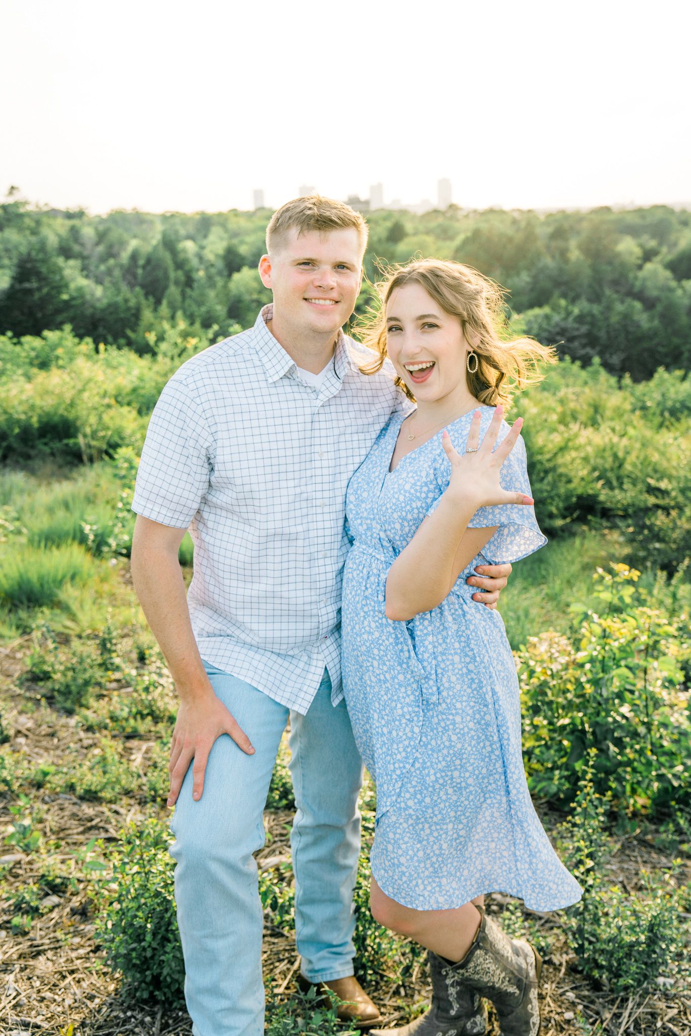 A couple poses outdoors in coordinating blue and white outfits against a natural green landscape on a sunny day.