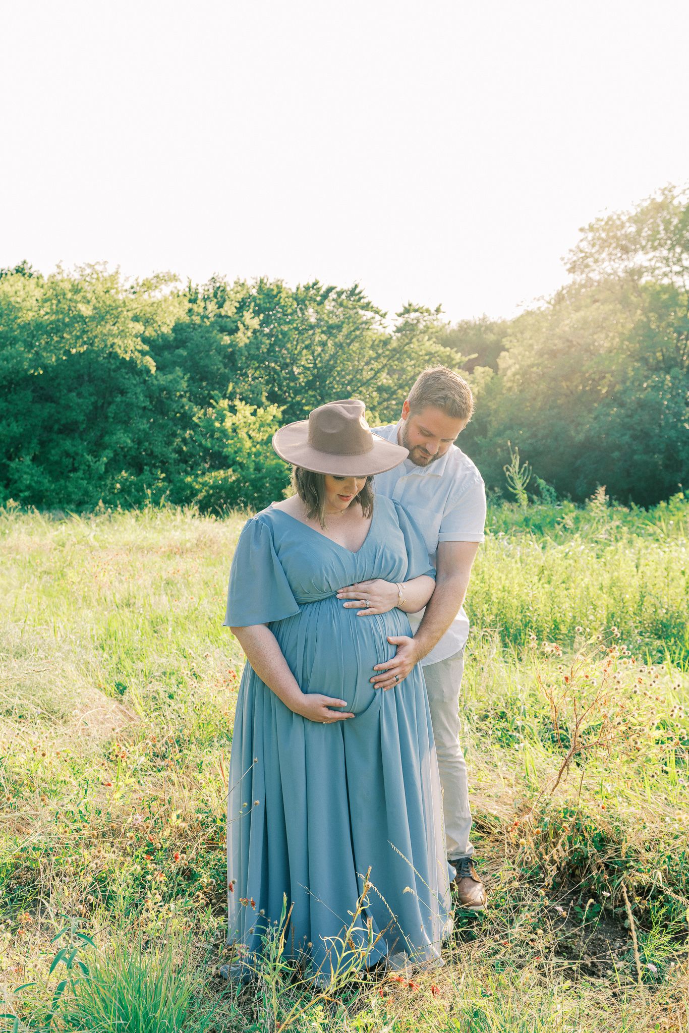 A couple in a sunlit meadow sharing a tender moment during a maternity photoshoot, with her wearing a flowing blue dress.