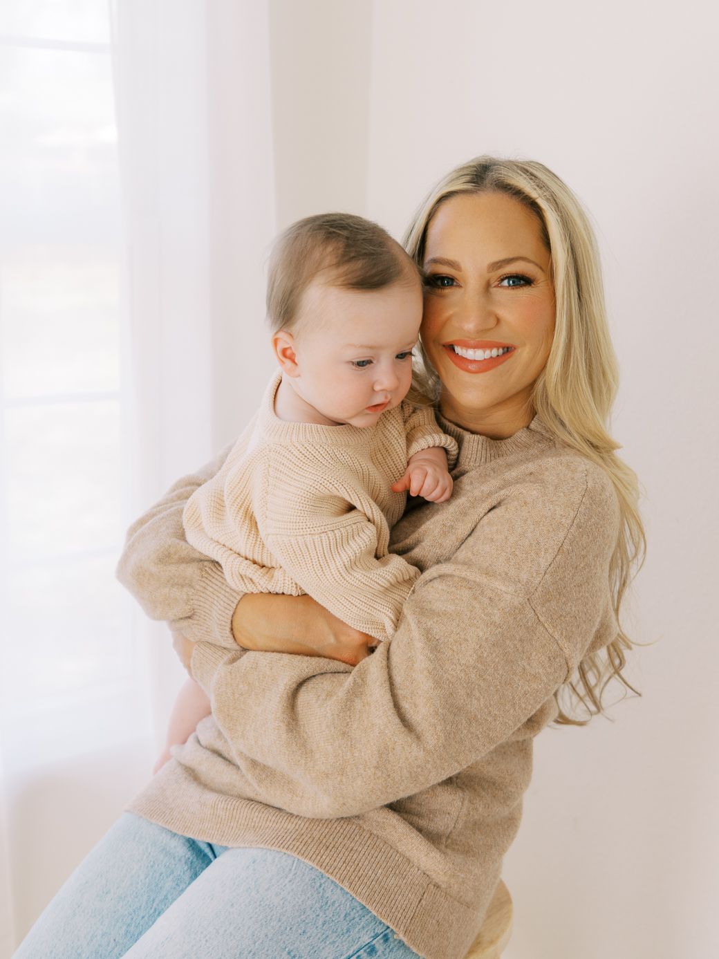 Mother and baby in cozy beige sweaters sharing warm embraces against bright white backdrop.