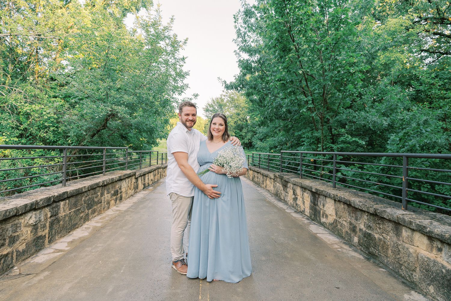 A couple stands embracing on a concrete bridge pathway with green foliage in the background.