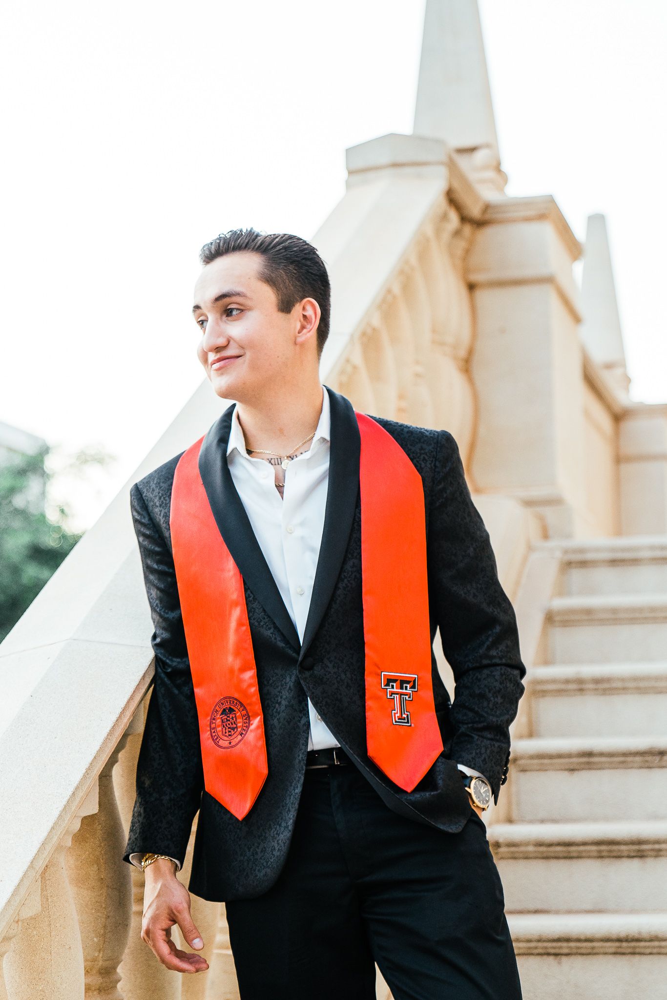 Graduate in black suit and red stole smiles while sitting on decorative stone staircase.