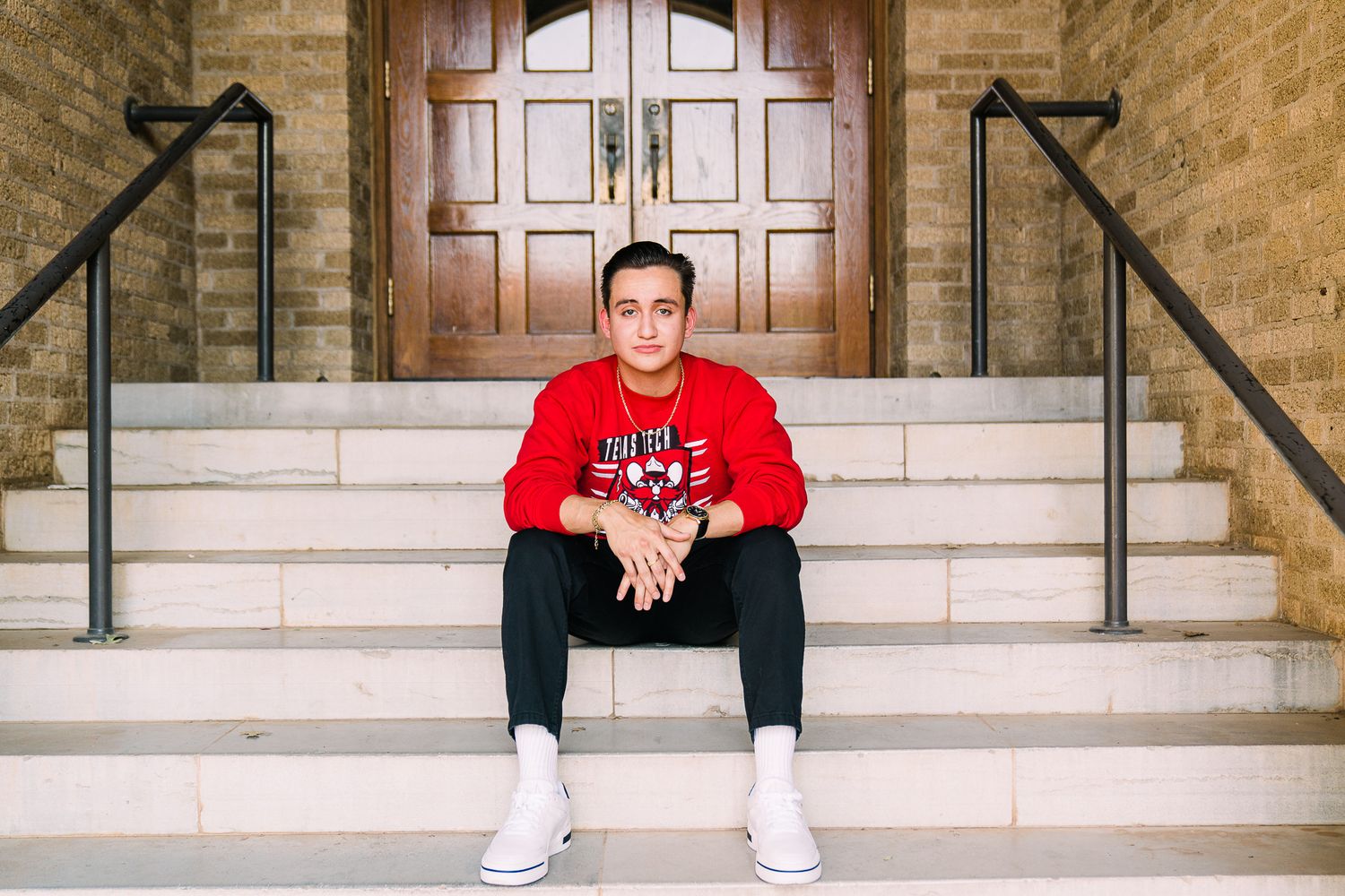 Person in red sweatshirt and white sneakers sits on concrete steps in front of brown brick wall and wooden doors.