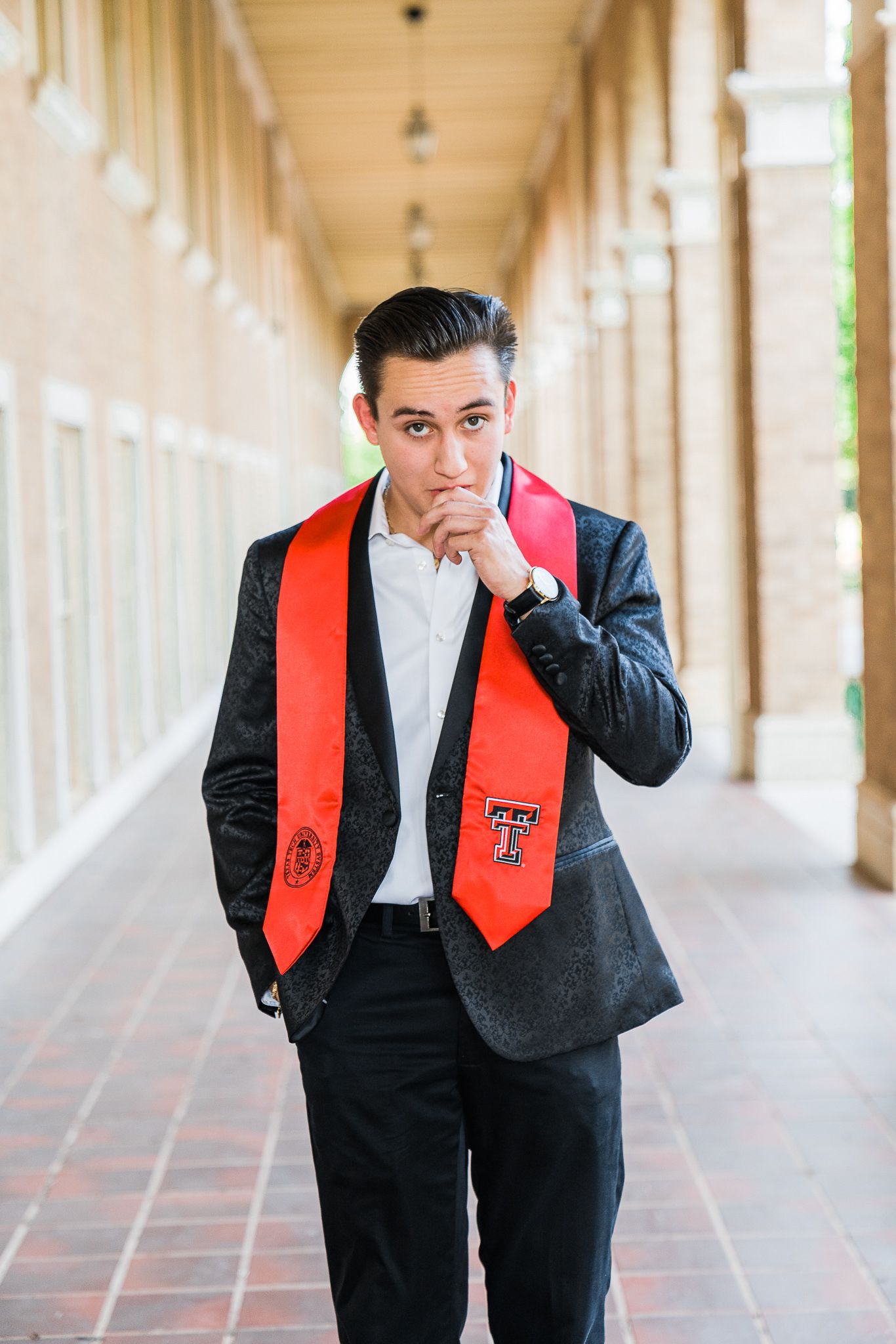 Graduate in formal attire with red graduation stole posing thoughtfully in a covered walkway.