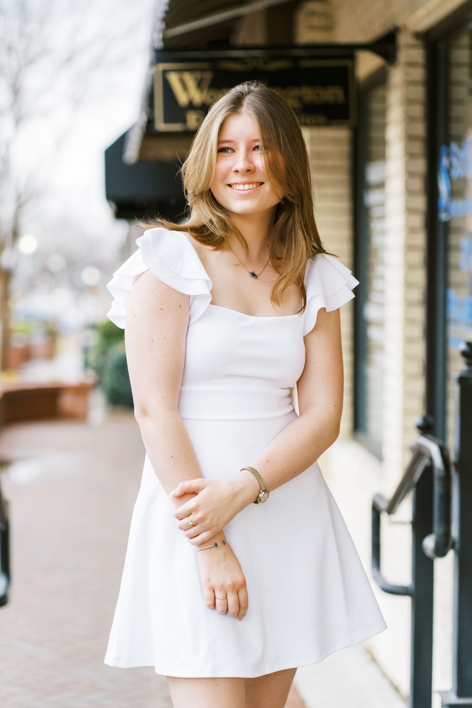 A person wearing a white ruffled dress poses outdoors near a storefront.