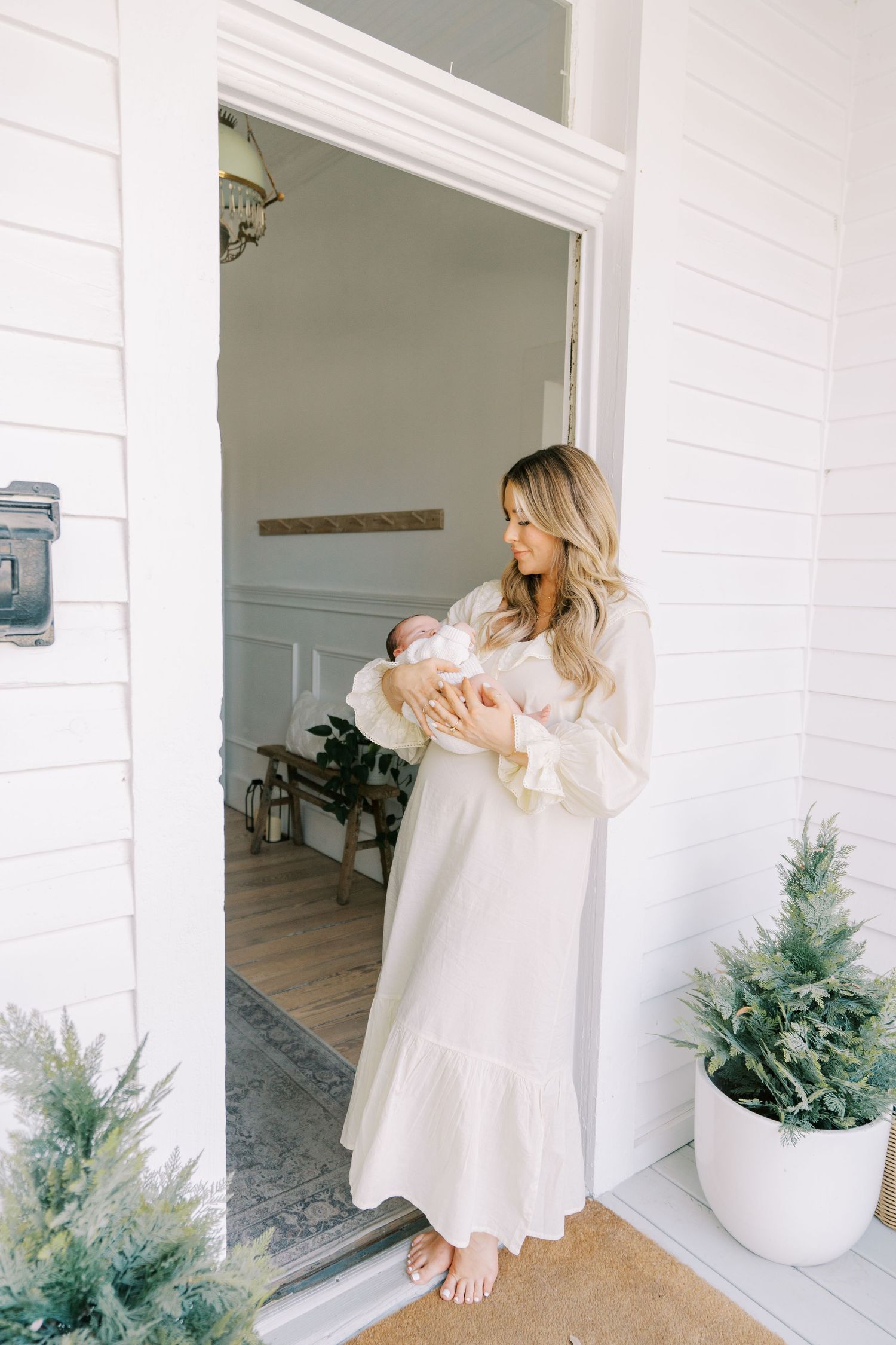 Woman in flowing white dress cradles newborn while standing by white columns and potted greenery.
