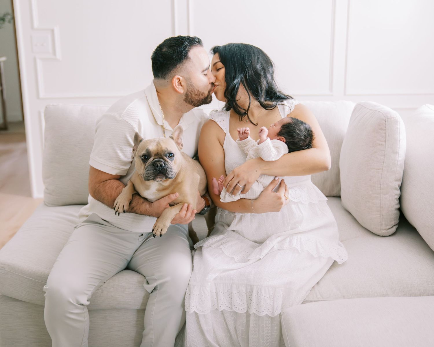 Couple kissing on white sofa with French Bulldog and newborn baby in bright living room.