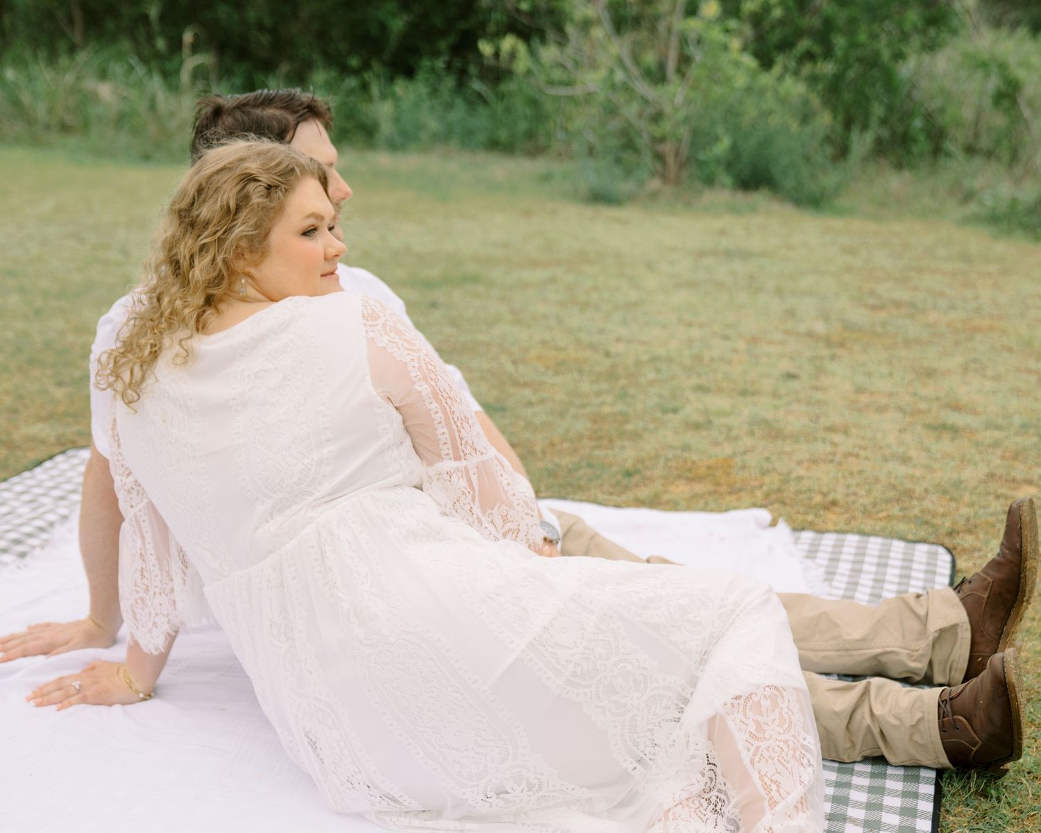 Couple relaxing on checkered picnic blanket outdoors, woman in white lace dress and brown boots.