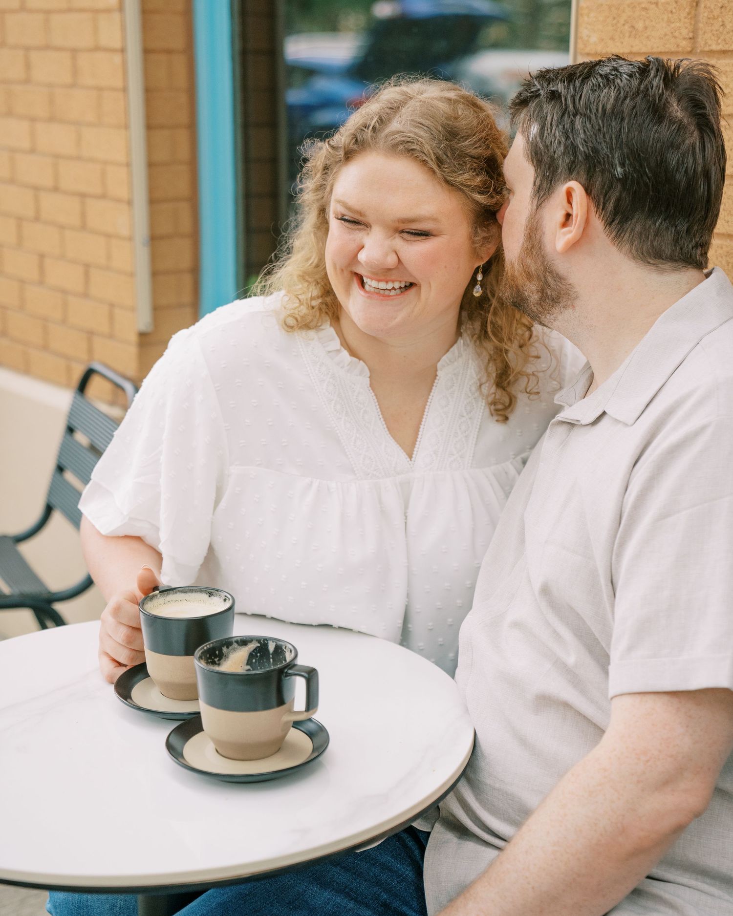A woman laughs joyfully while sitting with her partner at an outdoor cafe table with coffee mugs.