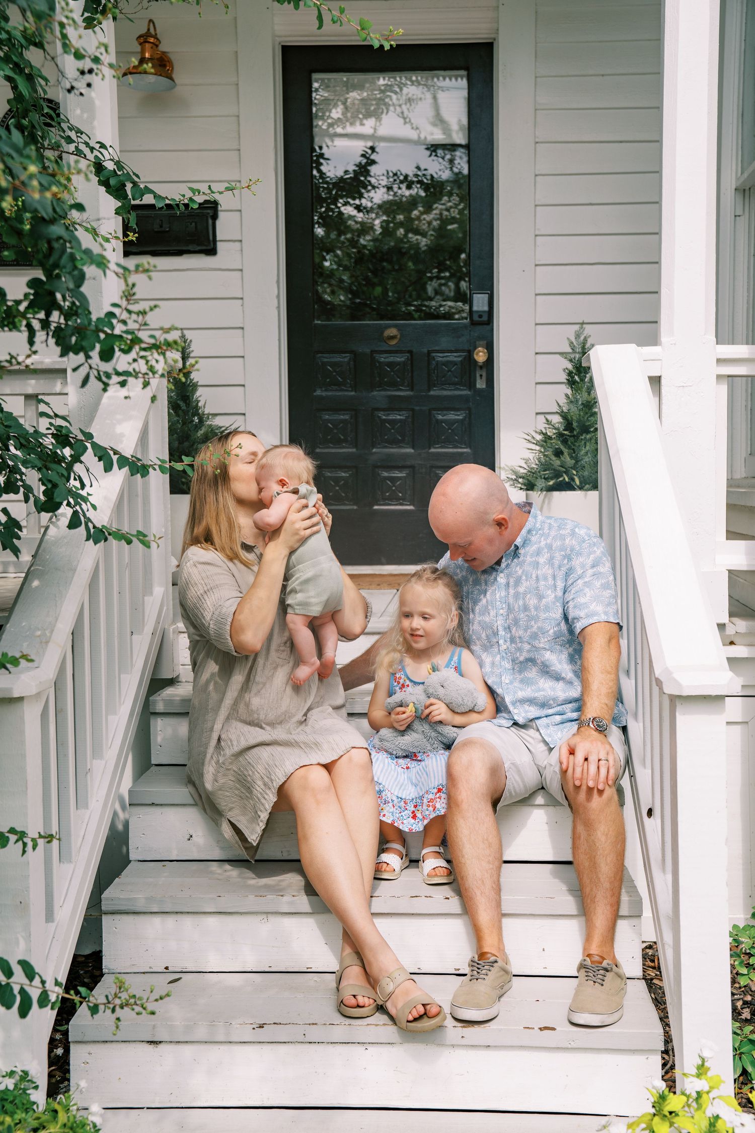 Family of four sitting on white porch steps, parents holding two young children outside a home with a dark front door.