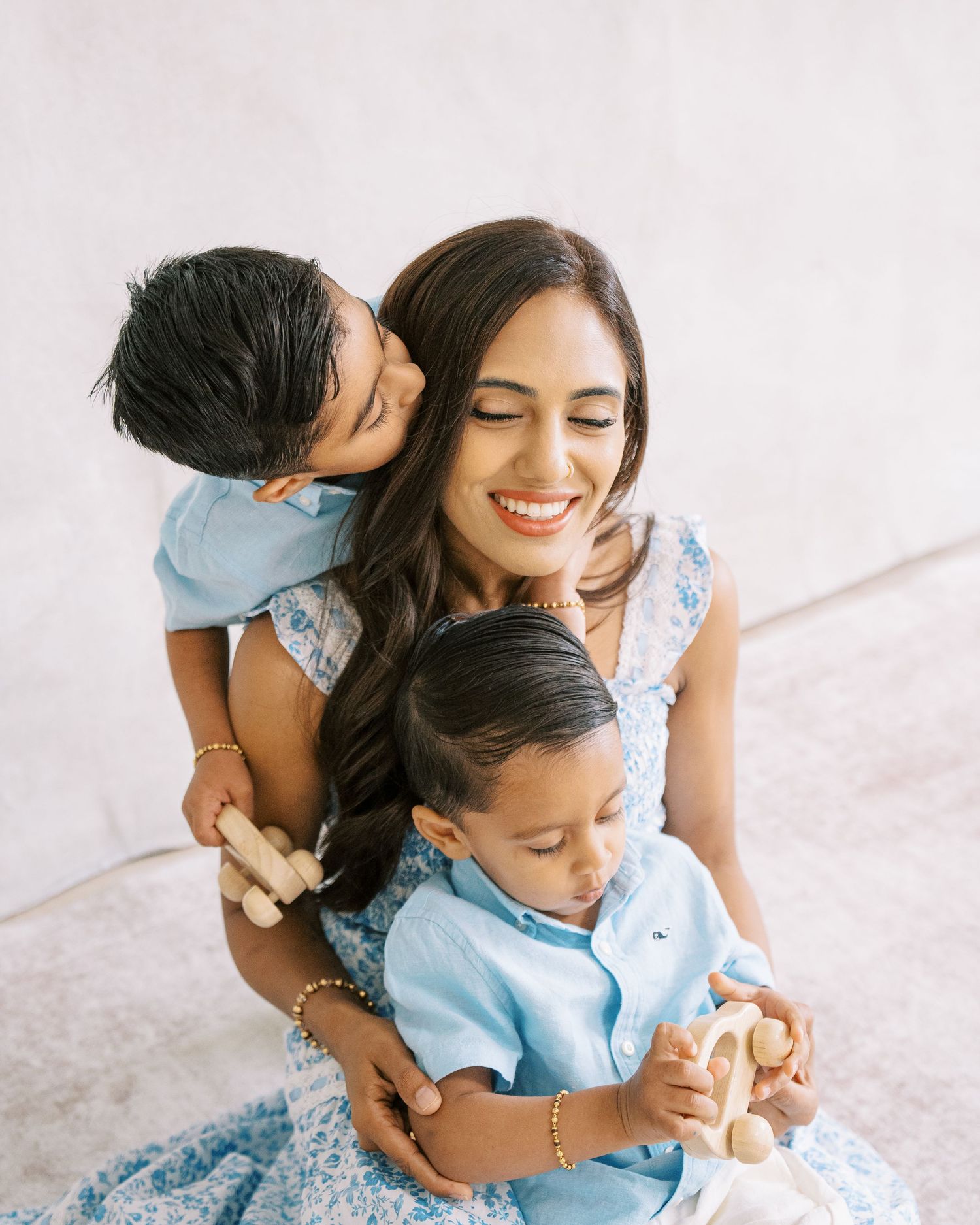 A smiling woman in a blue floral dress hugs two young boys in light blue outfits holding wooden toys.