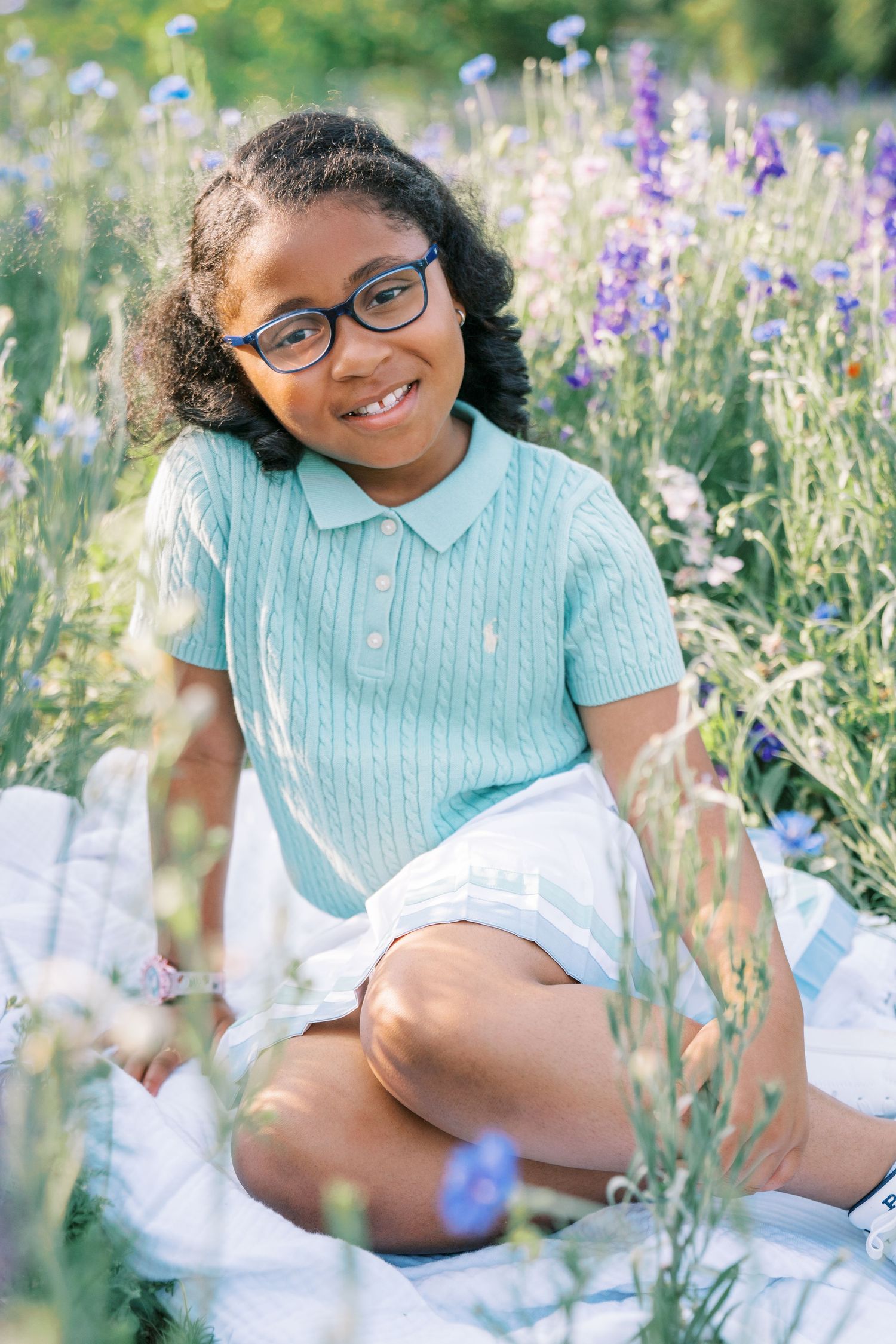 A smiling young girl wearing glasses and a teal polo shirt sits in a colorful wildflower field on a white blanket.