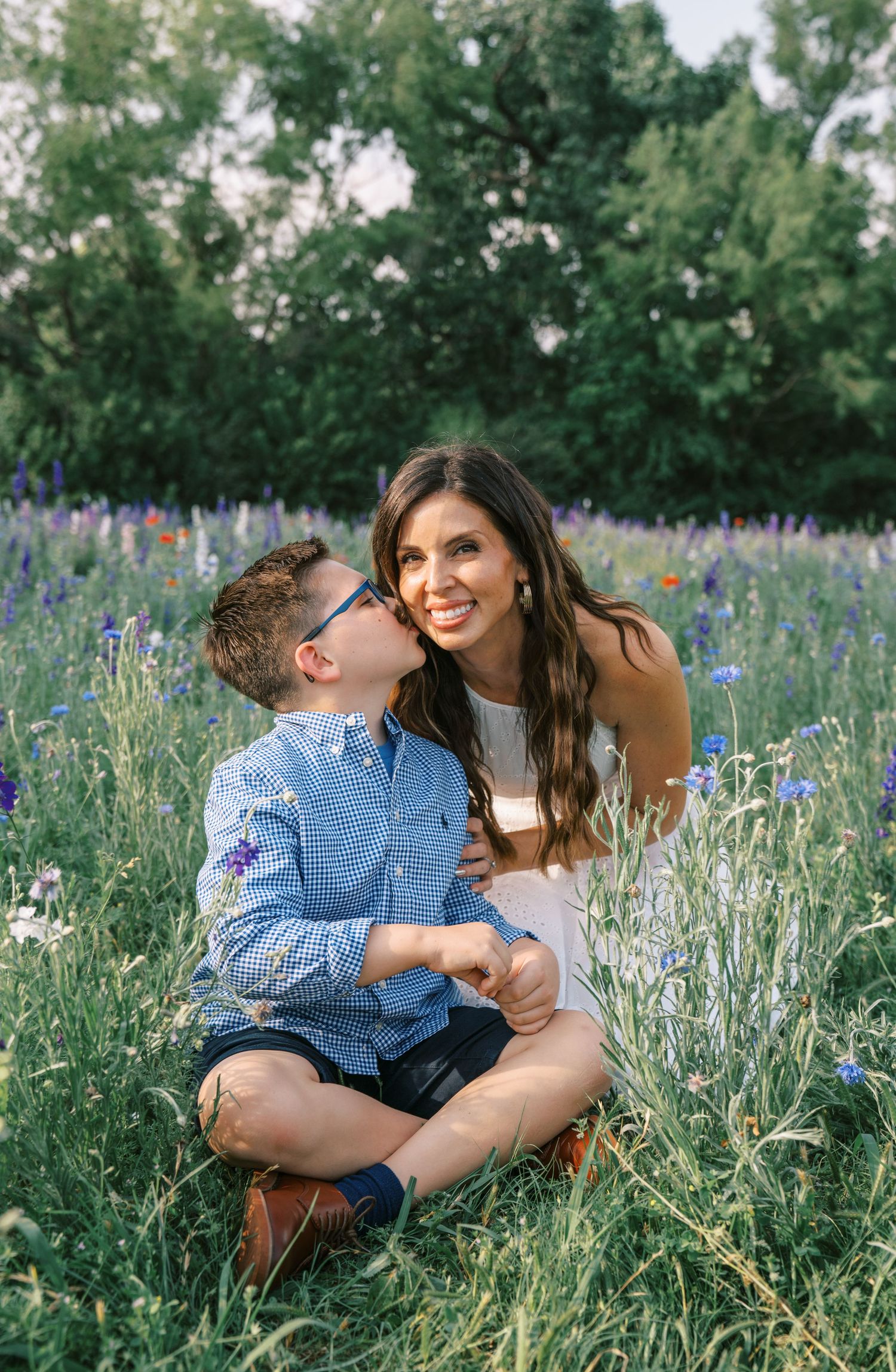 A young boy kisses his smiling mother on the cheek while sitting together in a vibrant wildflower field.