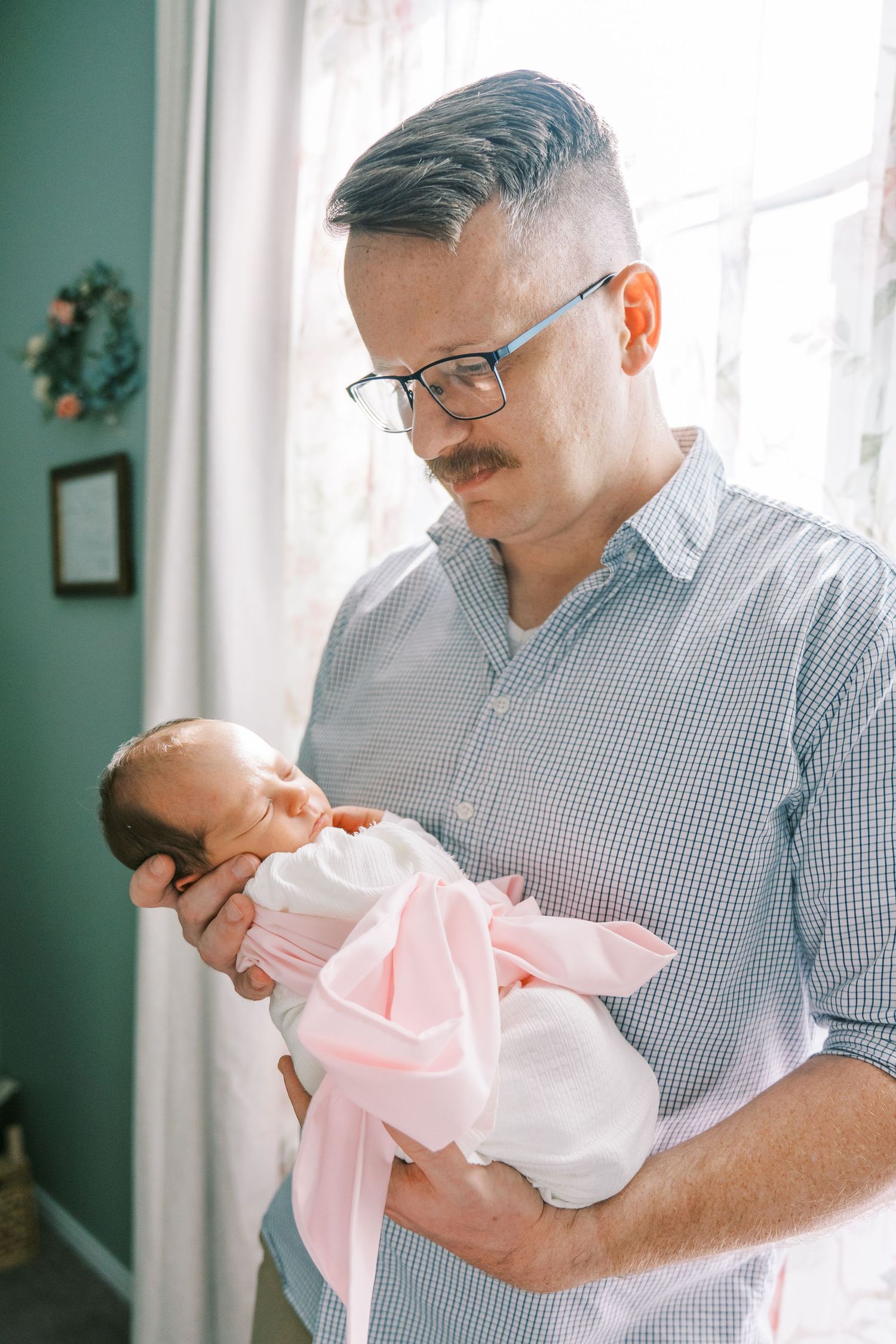 A tender moment between a parent in a striped shirt and glasses holding a sleeping newborn baby in pink and white.