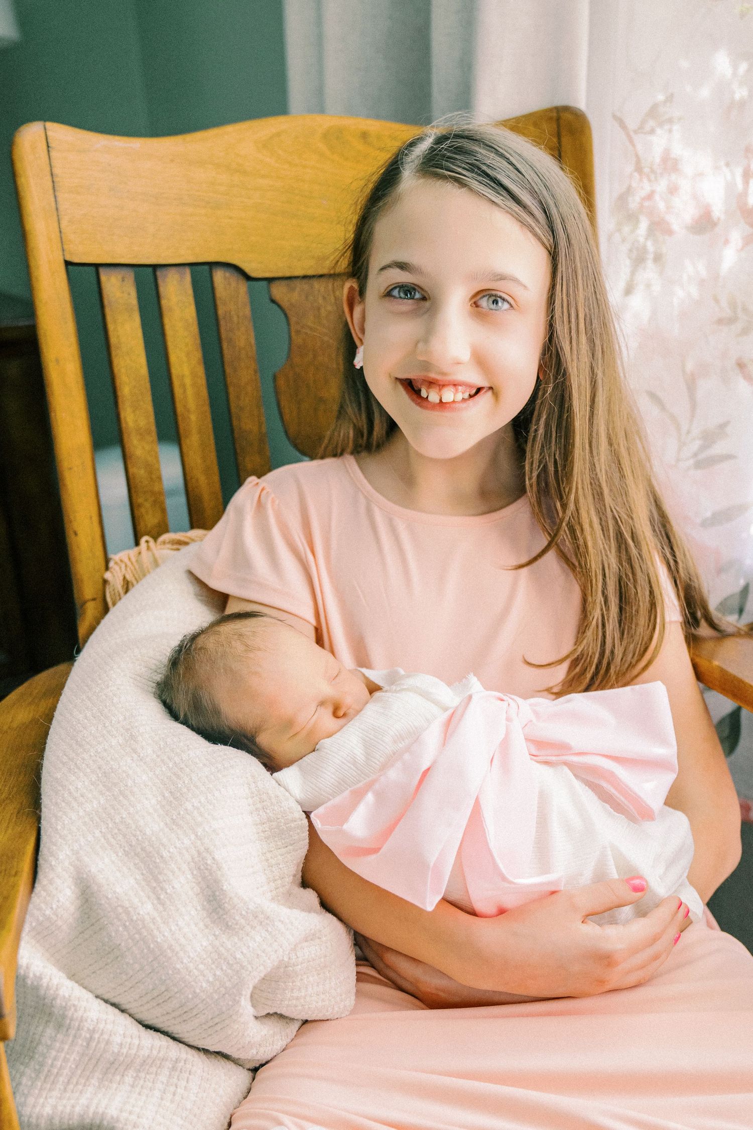 Young girl in pink dress holding newborn sibling while sitting in wooden chair.